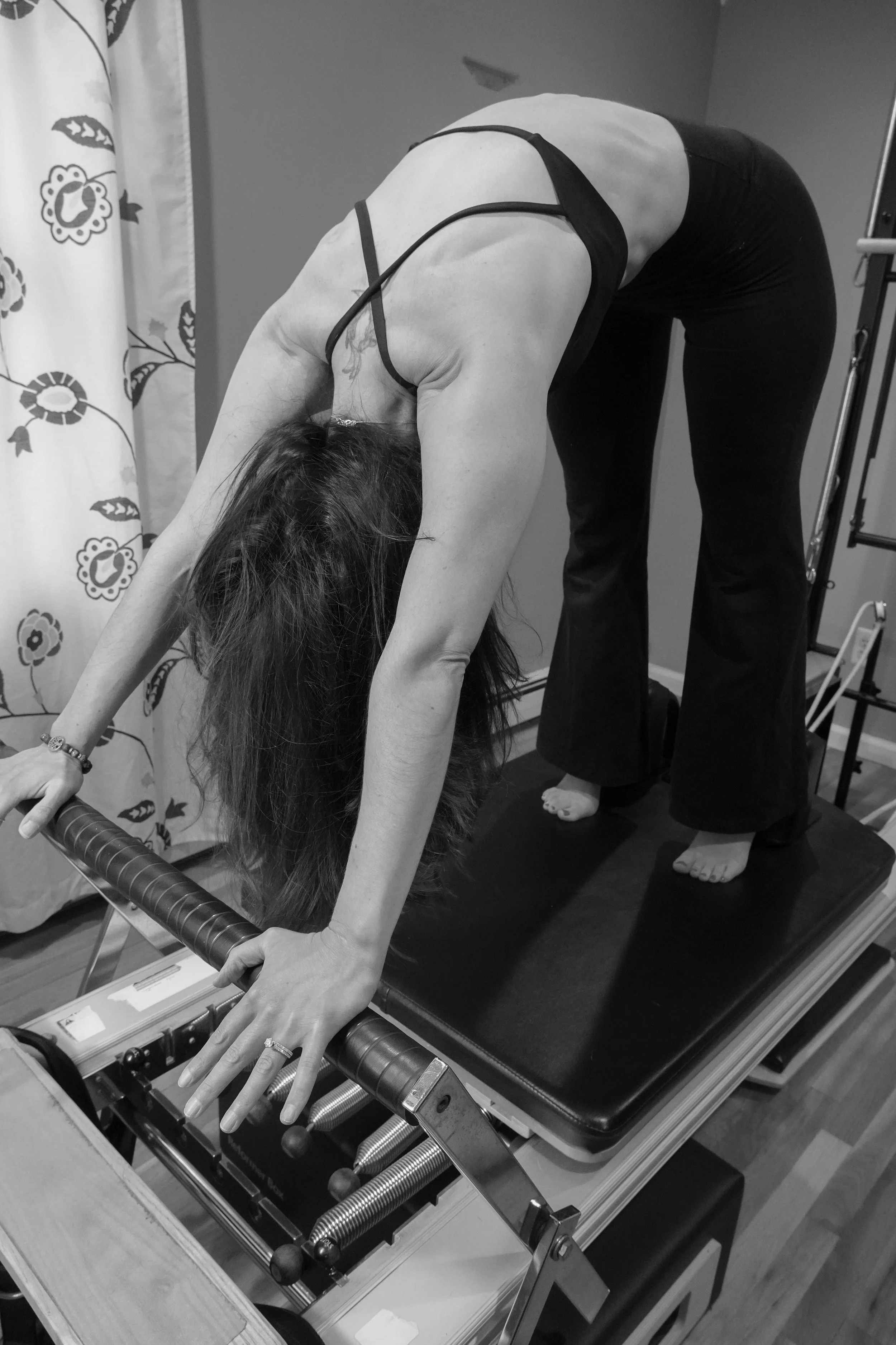 A woman in black clothes is bending forward with her hands on a foosball table, appearing to be playing or leaning over it, in a room with a patterned curtain and various equipment.