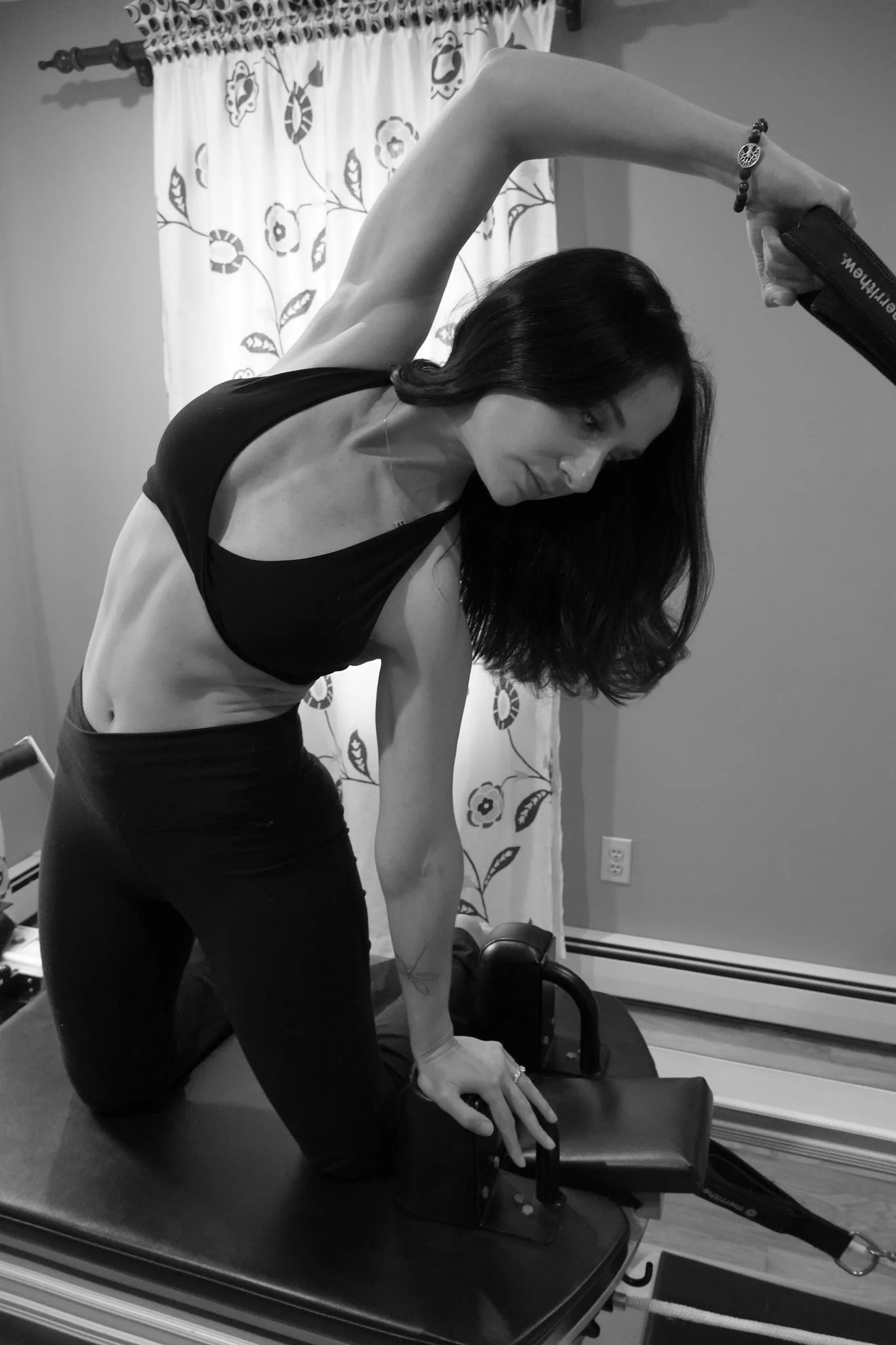 A woman practicing yoga or stretching on a reformer machine in a home gym or workout space. She is leaning forward with one arm extended overhead and the other arm resting on the reformer. She has long dark hair and is wearing a crop top and workout pants. There is a curtain with a butterfly and floral pattern in the background and exercise equipment around her.