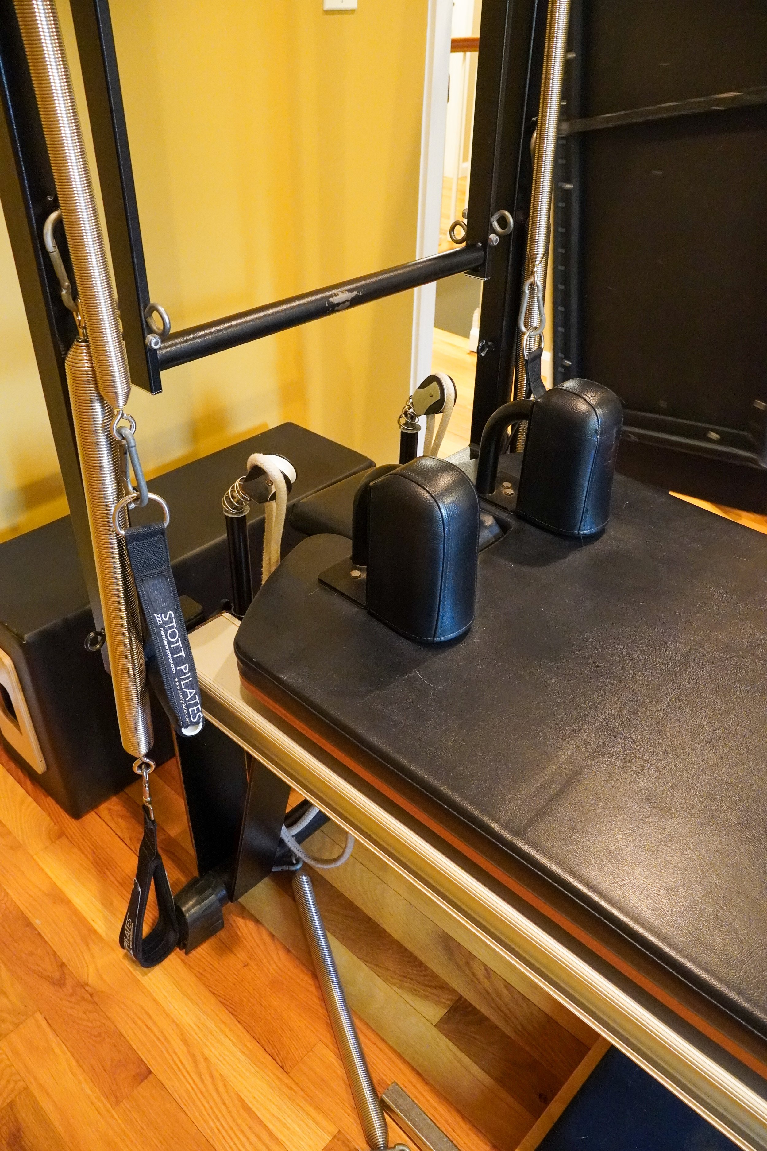 A Pilates reformer machine with a padded platform, adjustable springs, straps, and a bar, positioned in a room with a wooden floor and a beige wall.