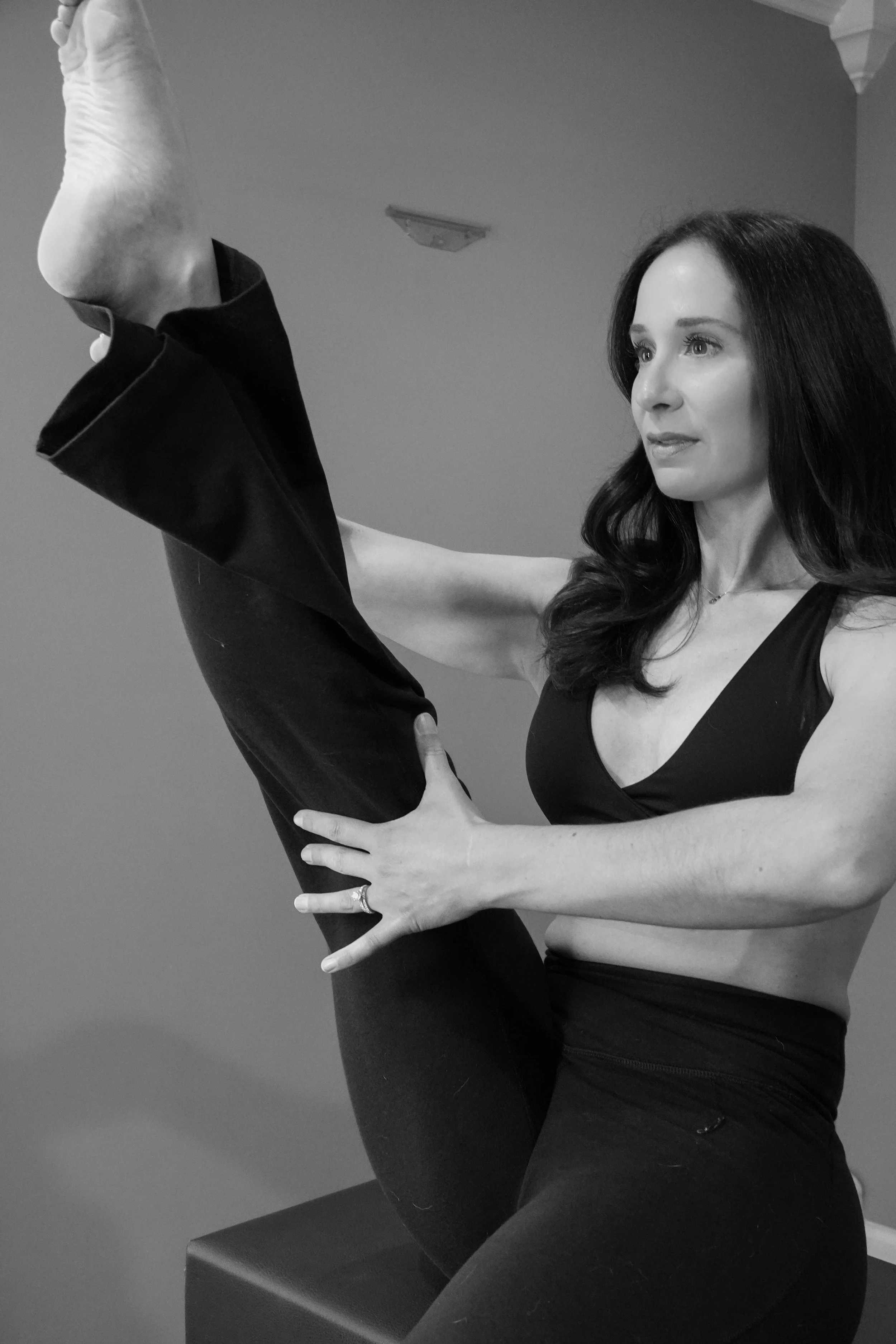 A woman with dark hair and light skin wearing a black dress and sitting on a bench, holding her raised leg with her hands. Her leg is bent, with her foot near her face, and she is looking at it. The image is in black and white.