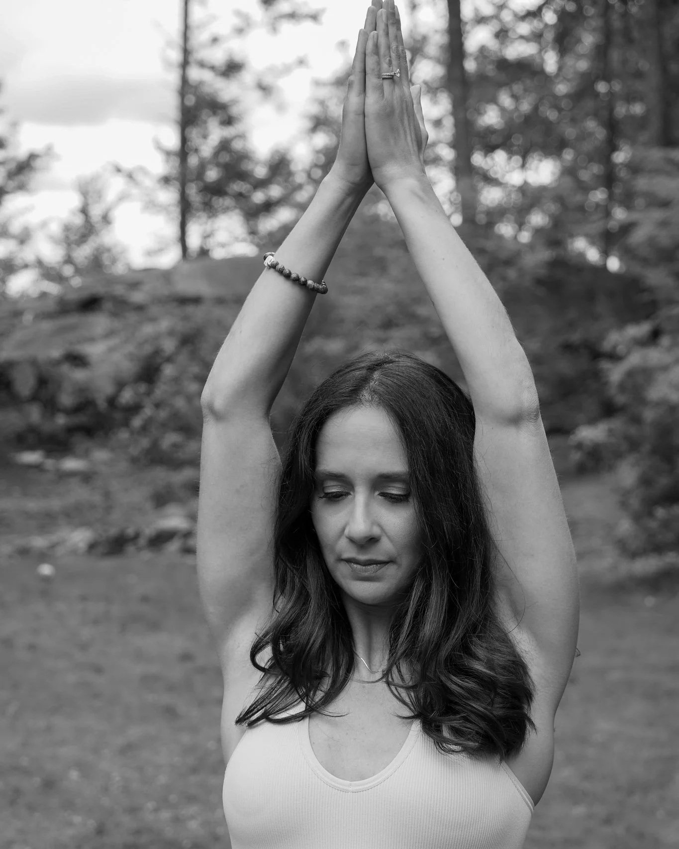 A woman with long wavy hair practicing yoga outdoors with her arms raised above her head and palms pressed together, eyes closed, in a forest setting.