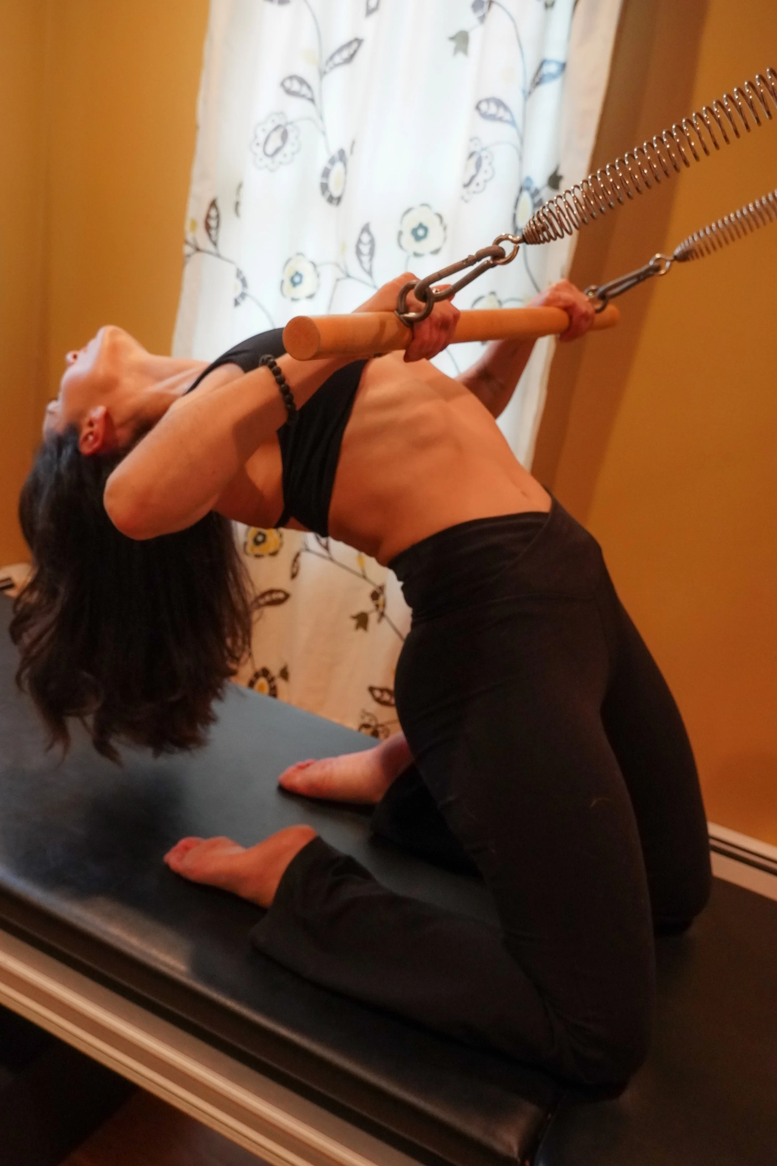 A woman practicing yoga on a black mat indoors, holding a yoga pose with a wooden massage bar connected to resistance bands, with a floral curtain and yellow walls in the background.