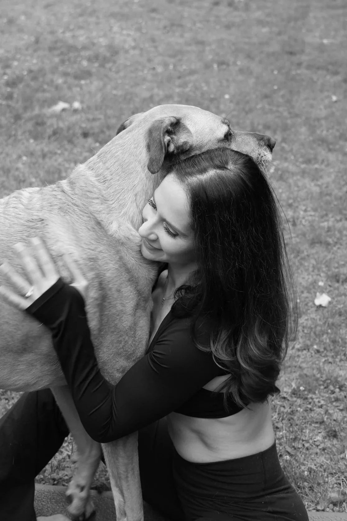 A woman hugging a large dog, both smiling, outdoors on a grassy area.