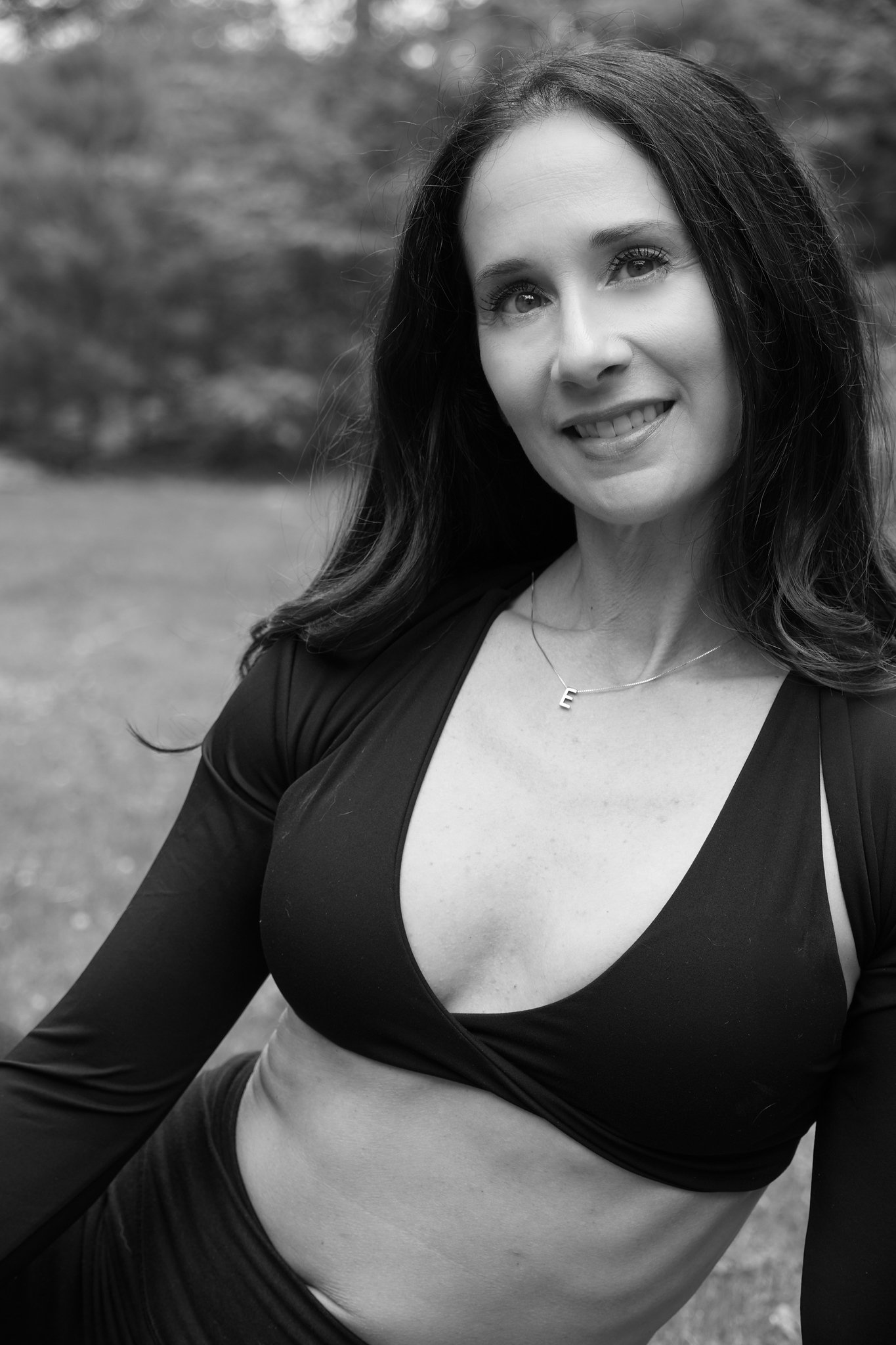 A black and white photo of a woman with dark hair wearing a black top with a deep neckline and a necklace with an 'E' pendant, outdoors with a blurred background.