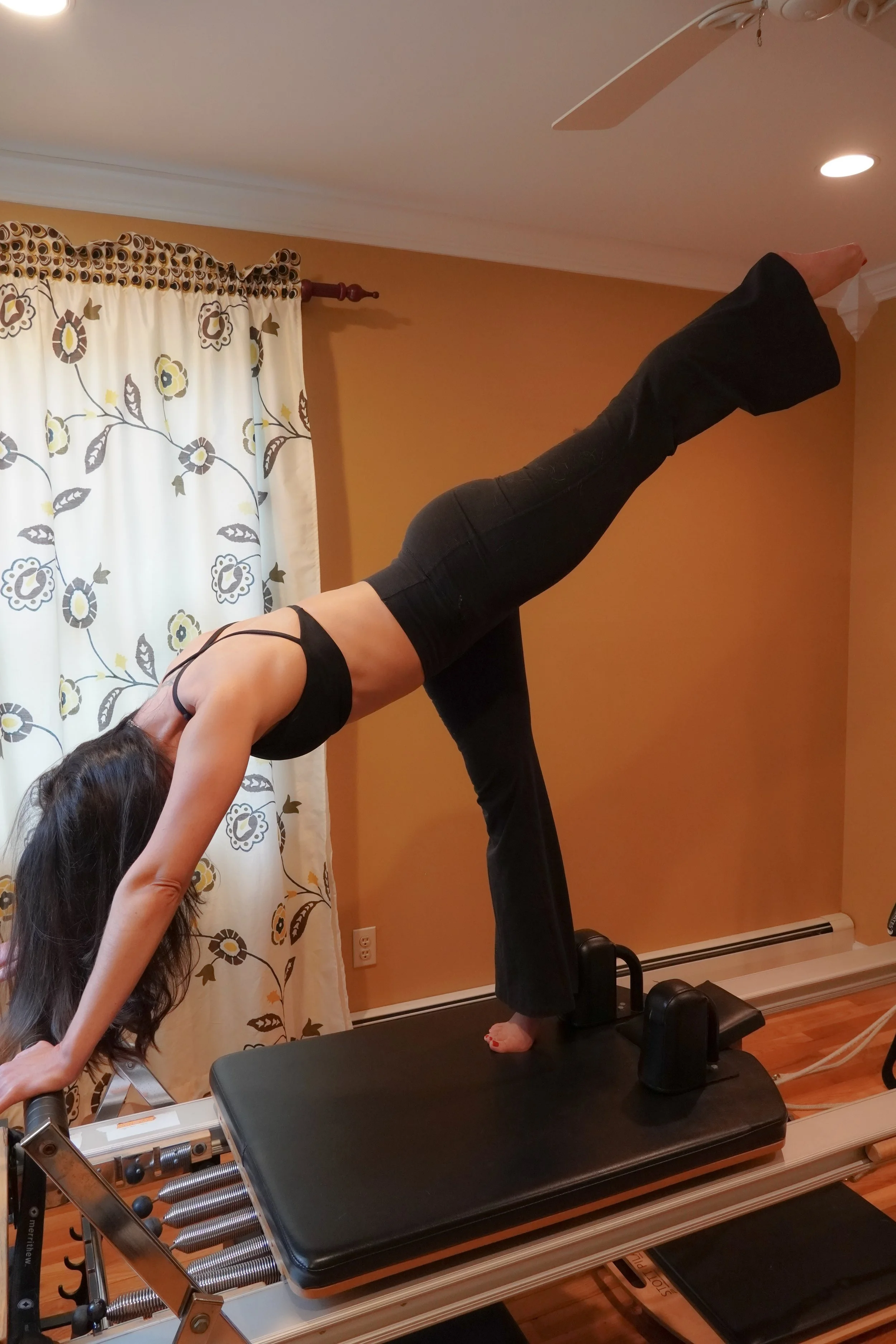 A woman practicing yoga on a reformer machine, performing a plank pose with her feet elevated on the wall, in a room with beige walls, floral curtains, and hardwood floors.
