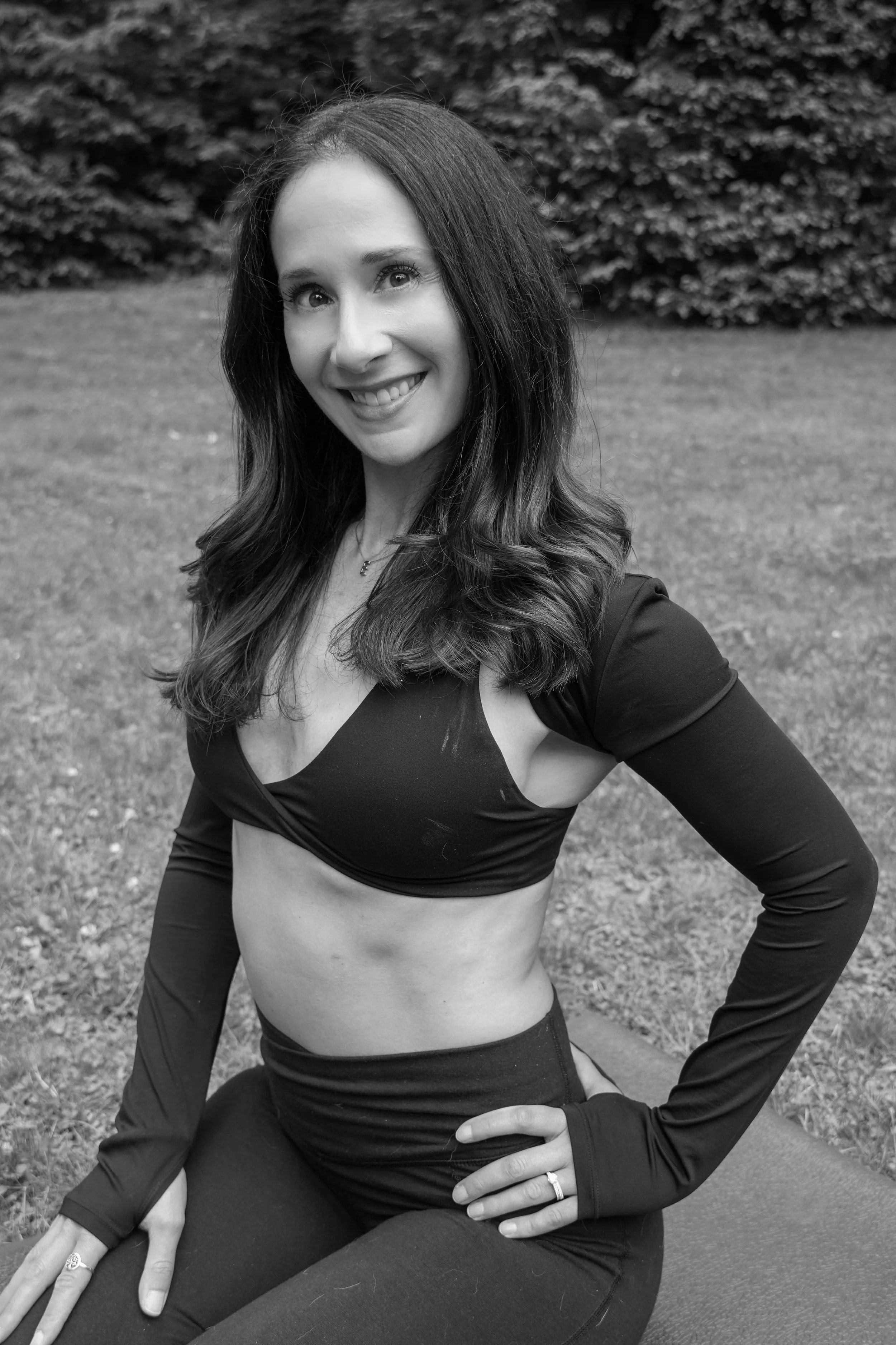 Black and white photo of a smiling woman with long hair, wearing a sports bra and leggings, sitting outdoors on the grass with her hand on her knee.