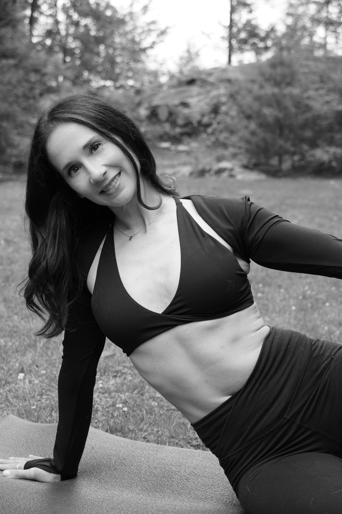 A woman with long dark hair is practicing yoga outdoors on a grass field, performing a side plank pose on a yoga mat. She is smiling and looks relaxed, with trees and foliage in the background.