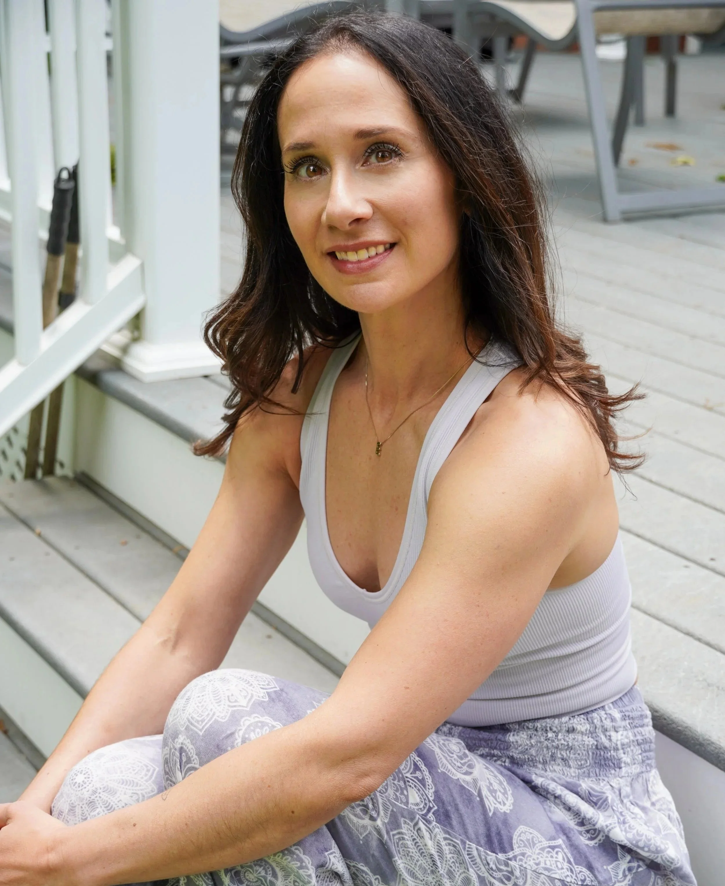A woman with long dark hair and brown eyes sitting outdoors on a wooden deck, smiling at the camera, wearing a white tank top and patterned white and gray pants.