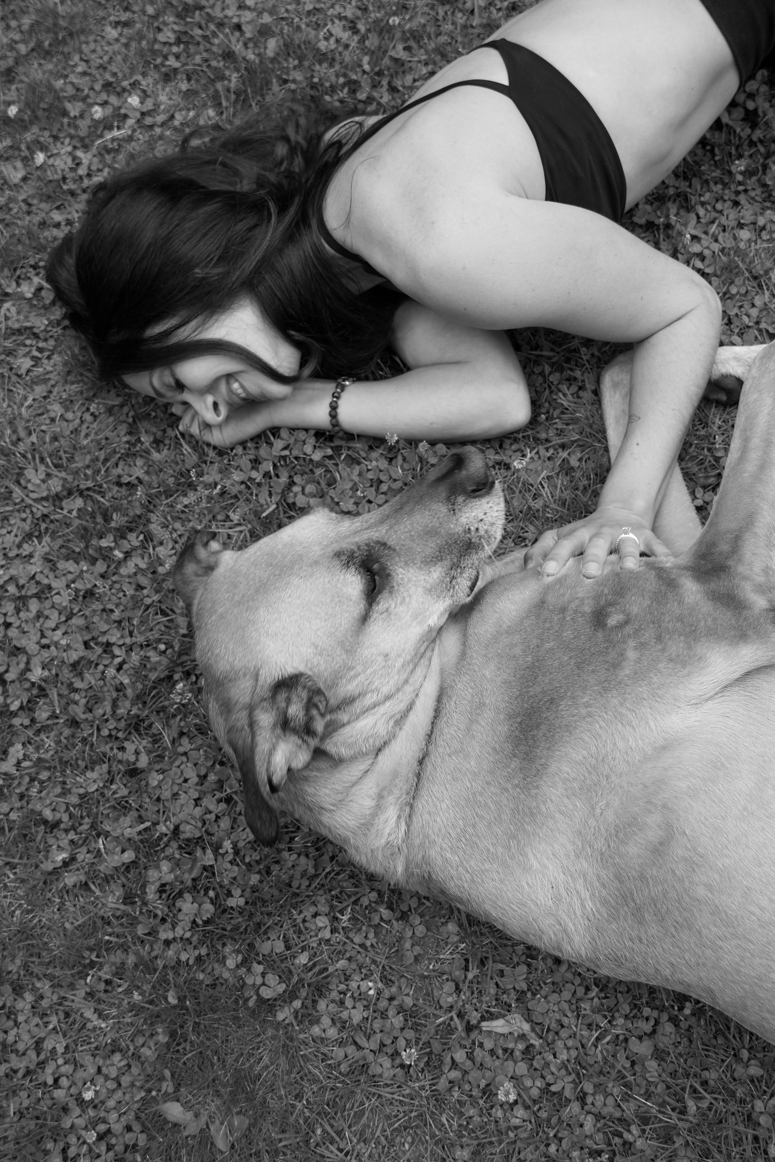 A woman lying on grass in black and white, smiling with her eyes closed, petting a large dog that is lying next to her, also appearing relaxed and content.