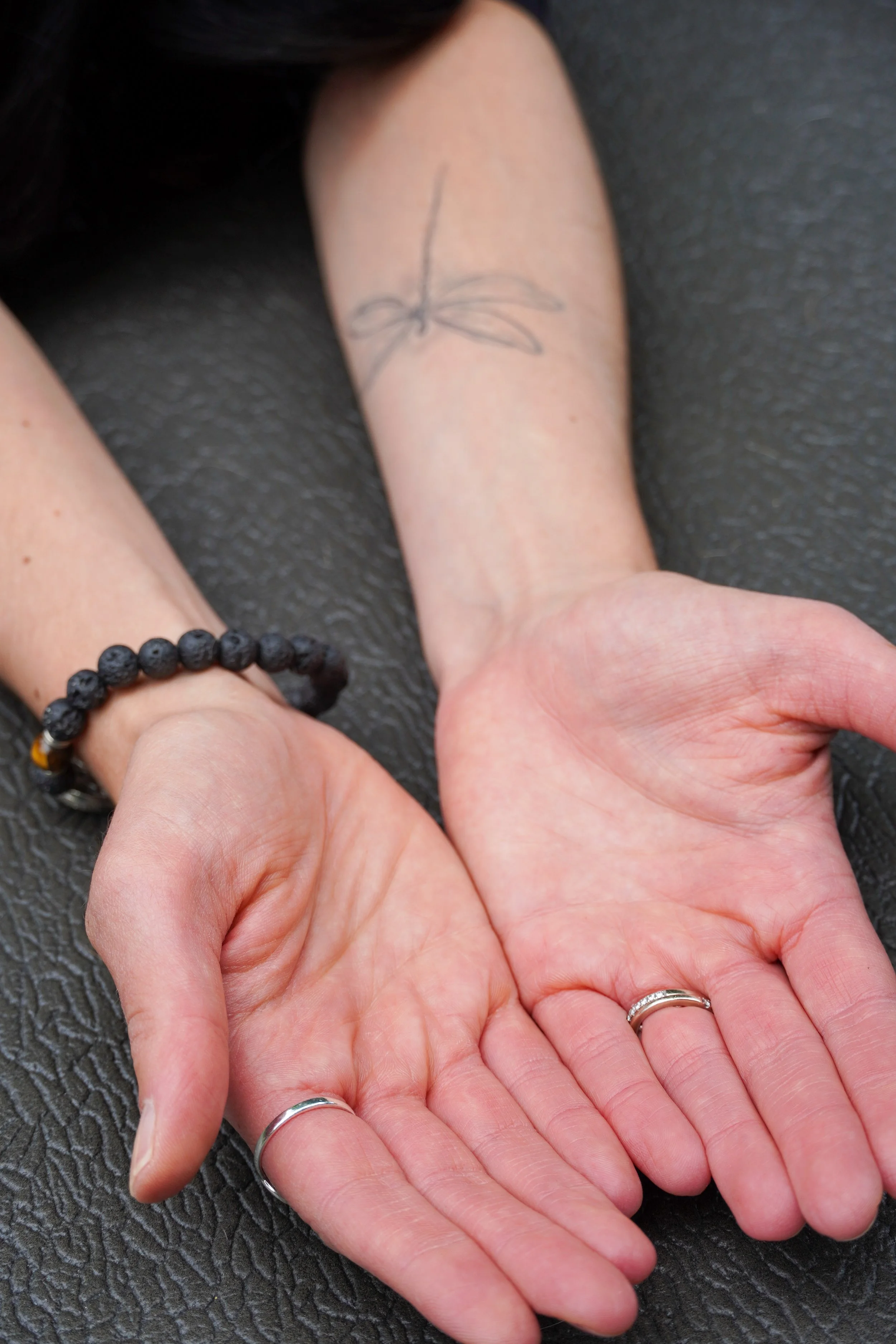 Close-up of two hands with wedding bands, one hand resting on a textured dark surface, the other hand slightly above with a wrist tattoo of a butterfly and a black beaded bracelet.
