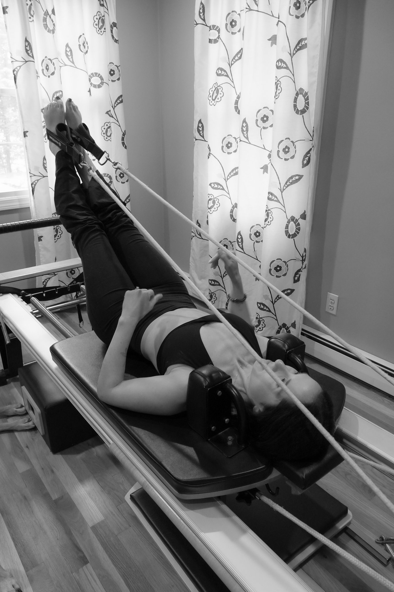 A person lying on a medical or therapeutic table with their legs elevated and secured by straps, in a room with curtains featuring a floral pattern, and a window with daylight outside.