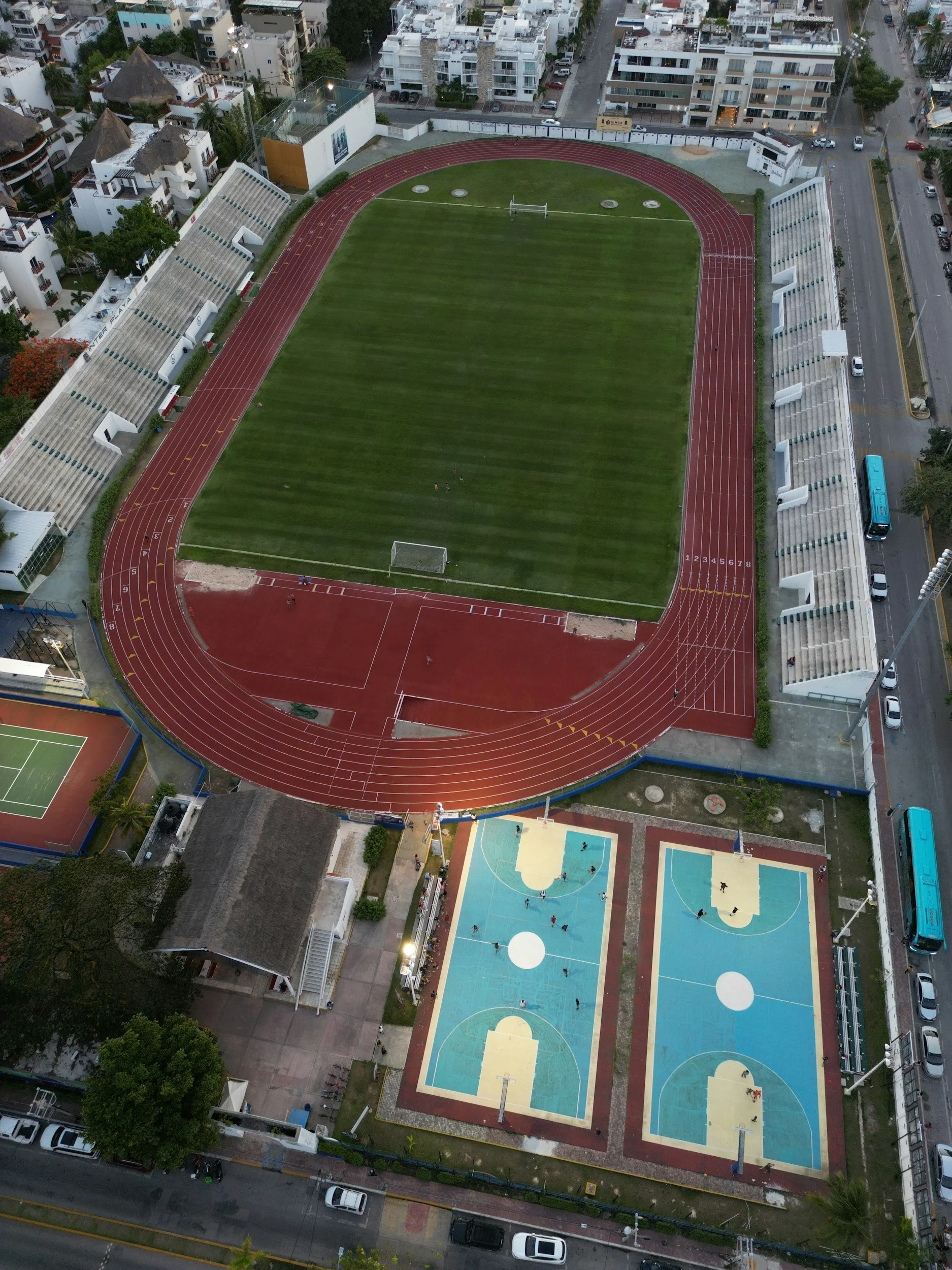 Aerial view of a sports complex with a soccer field, an athletics track, and basketball courts. Surrounding the complex are residential buildings and streets.