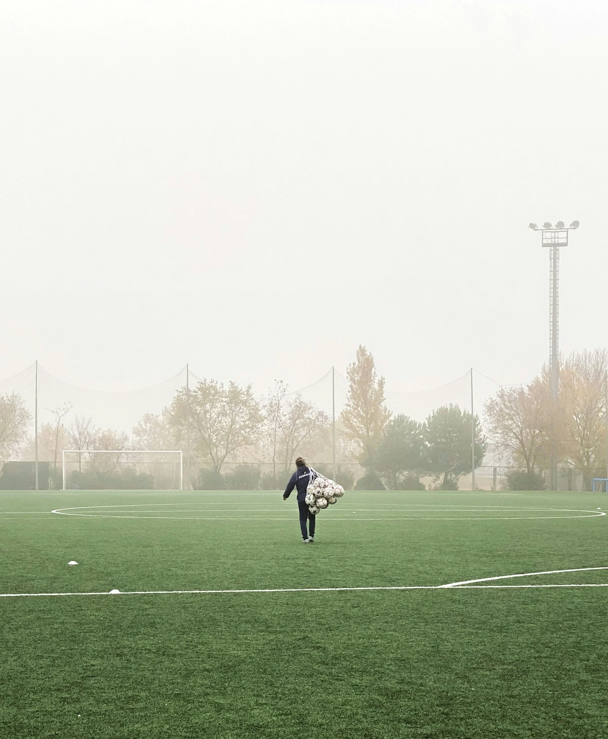 A person on a football field carrying a large pile of soccer balls in a net. The field is surrounded by trees and a tall light pole. The sky is foggy or cloudy.