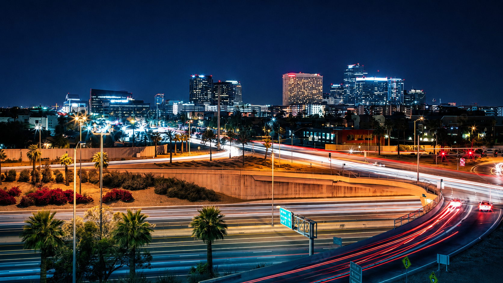 Nighttime cityscape with illuminated buildings, palm trees, and light trails from moving vehicles on highways and streets.