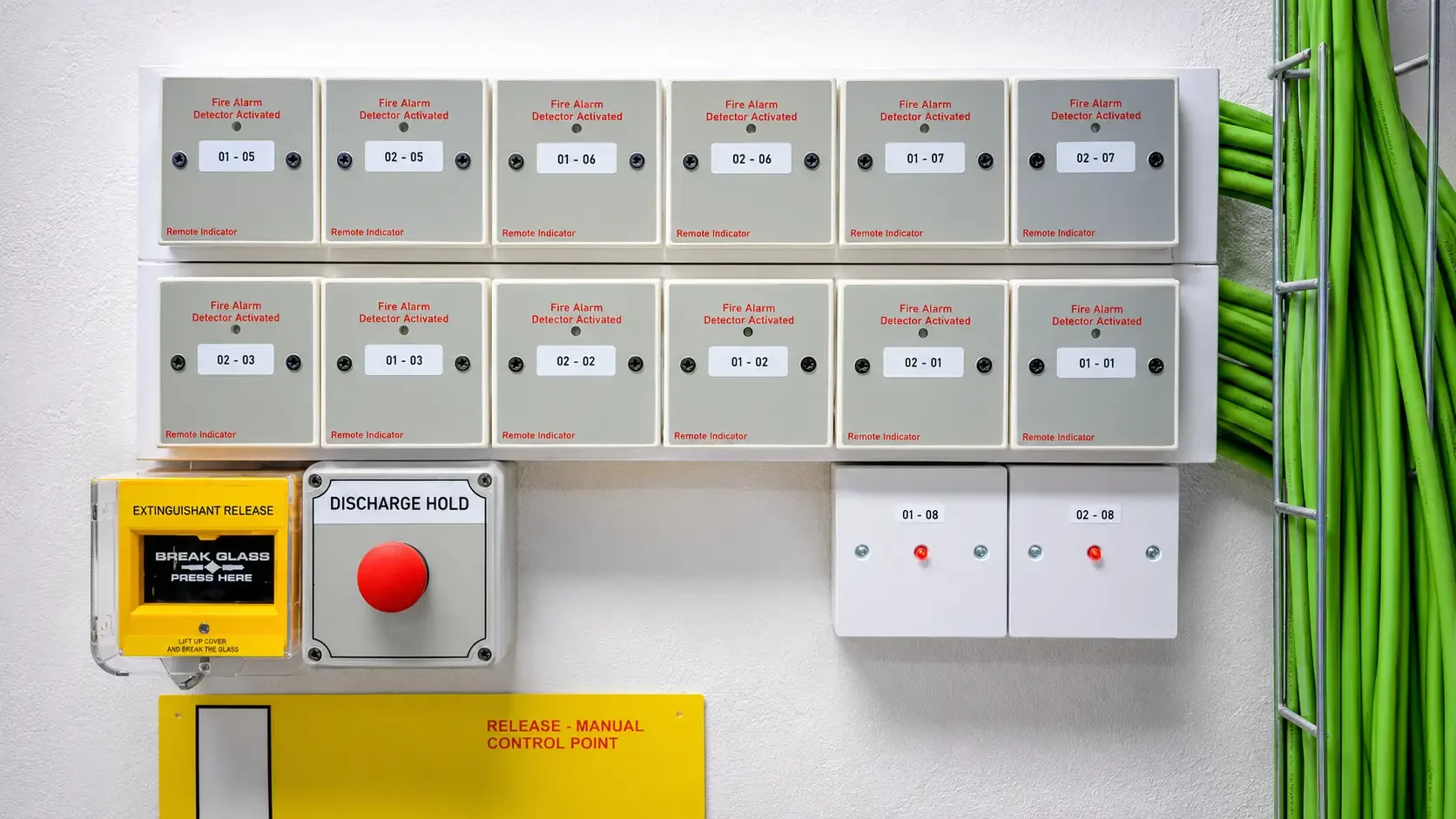 Fire alarm control panel with multiple alarm indicator boxes labeled with numbers, a yellow extinguishant release box, a discharge hold button, and some green pipes on the side.