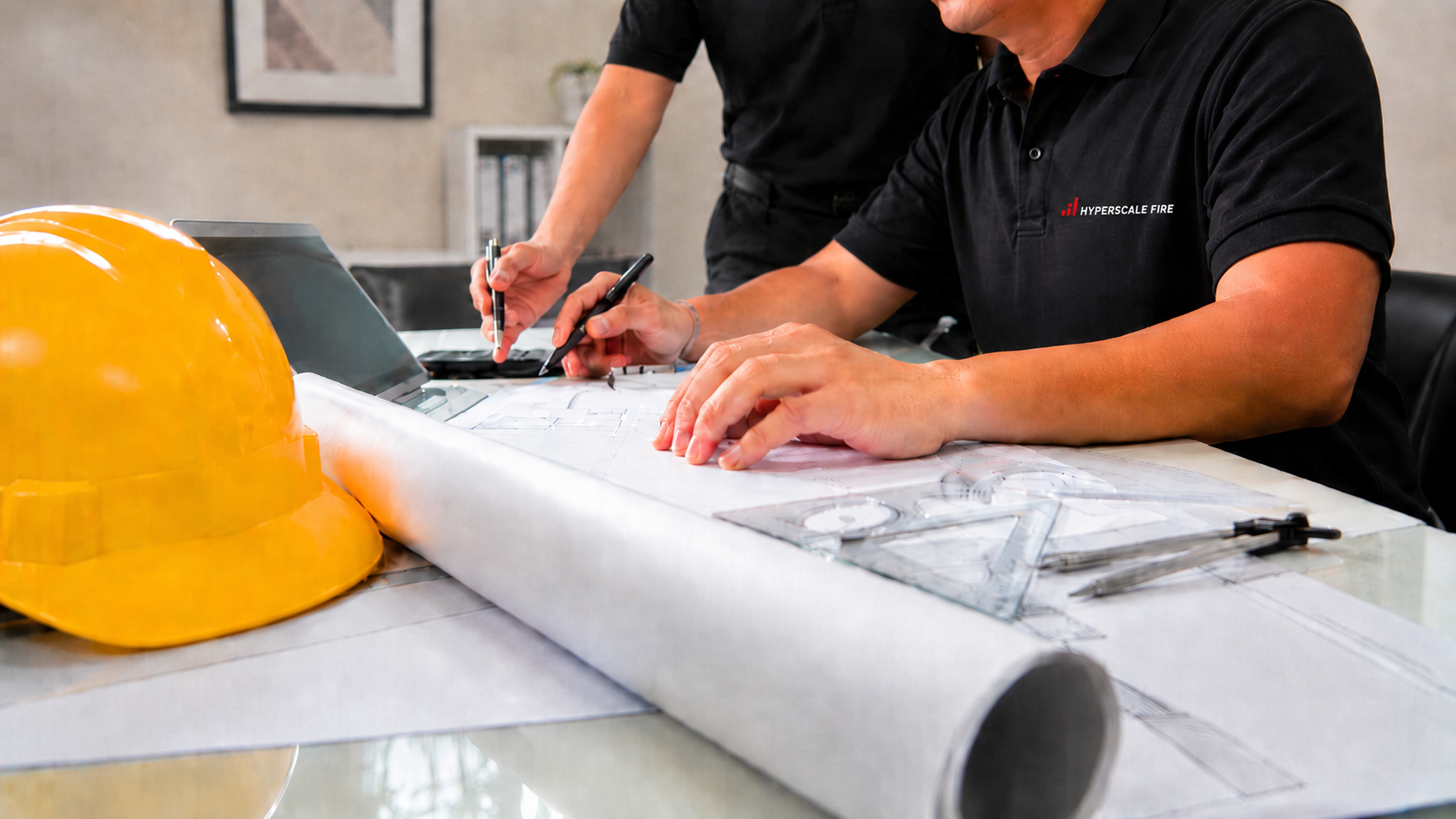 Two people working on architectural plans at a desk, with a yellow safety helmet and drafting tools, in an office setting.
