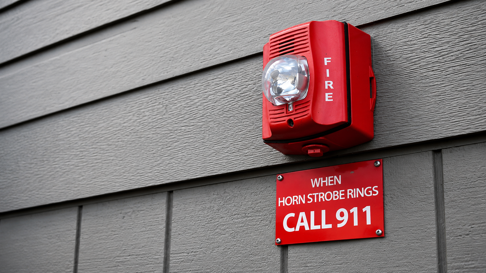 Red fire alarm with a clear dome lens mounted on a beige exterior wall with horizontal siding, below a red sign that reads, "When horn strobe rings call 911."