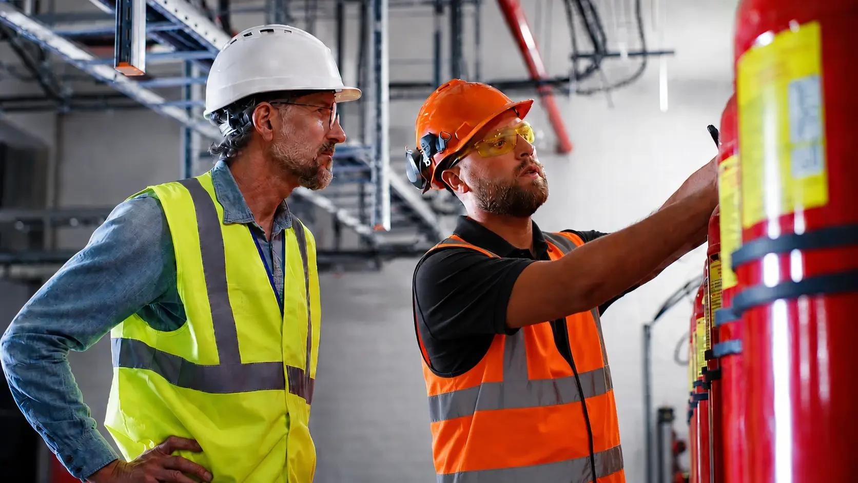 Two male workers in safety vests and hard hats inspecting fire extinguishers in an industrial setting.