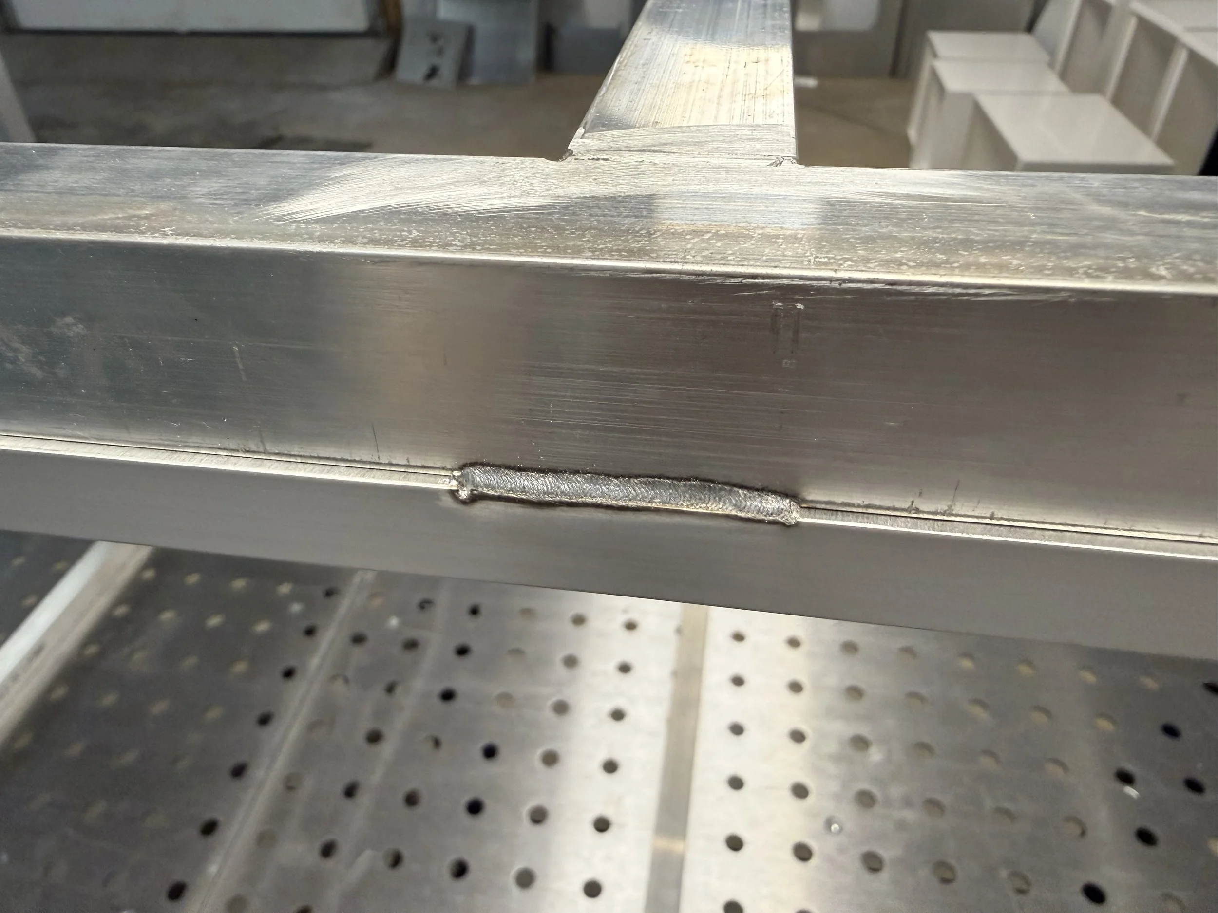 Close-up of a metallic cross-section of aluminum or steel with visible welding marks, placed on a metallic perforated surface.