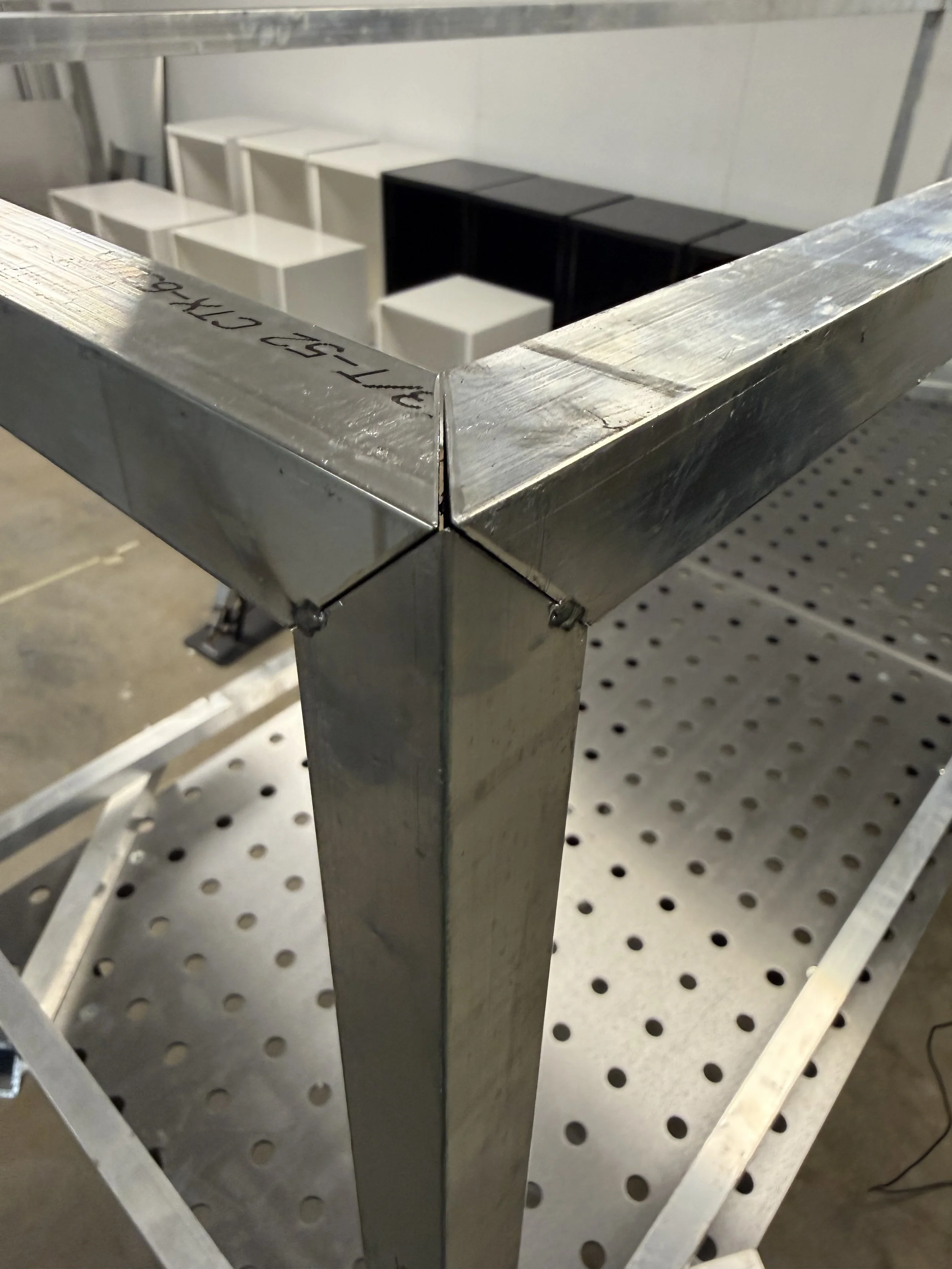 Close-up of a metallic corner of a shelving unit or table, with a white perforated metal surface beneath, and stacked white and black cube-shaped objects in the background.