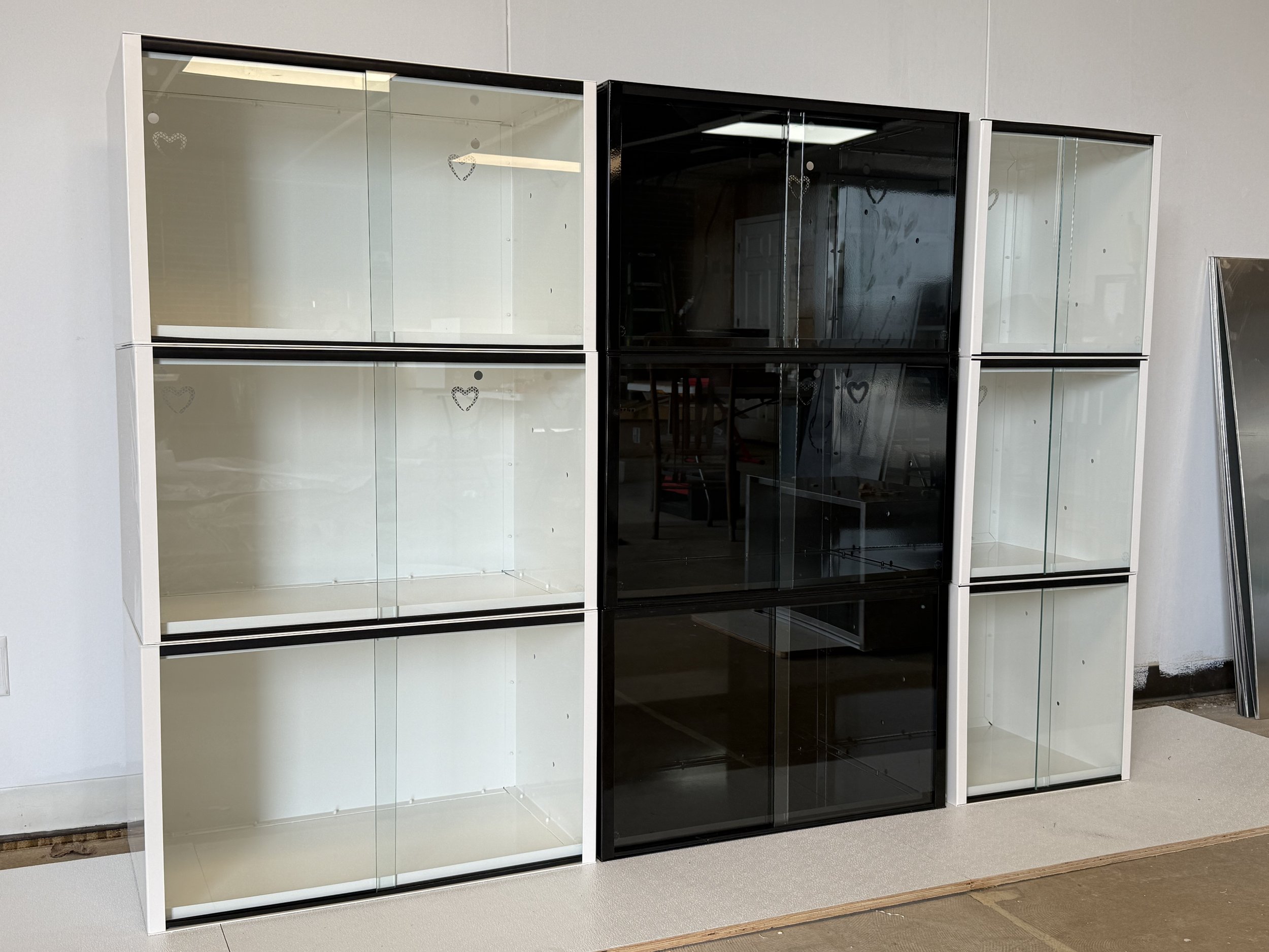Multiple black and white  powder coated aluminum arboreal reptile or amphibian enclosures, with heart shaped ventilation, are placed next to a wall in an indoor setting.
