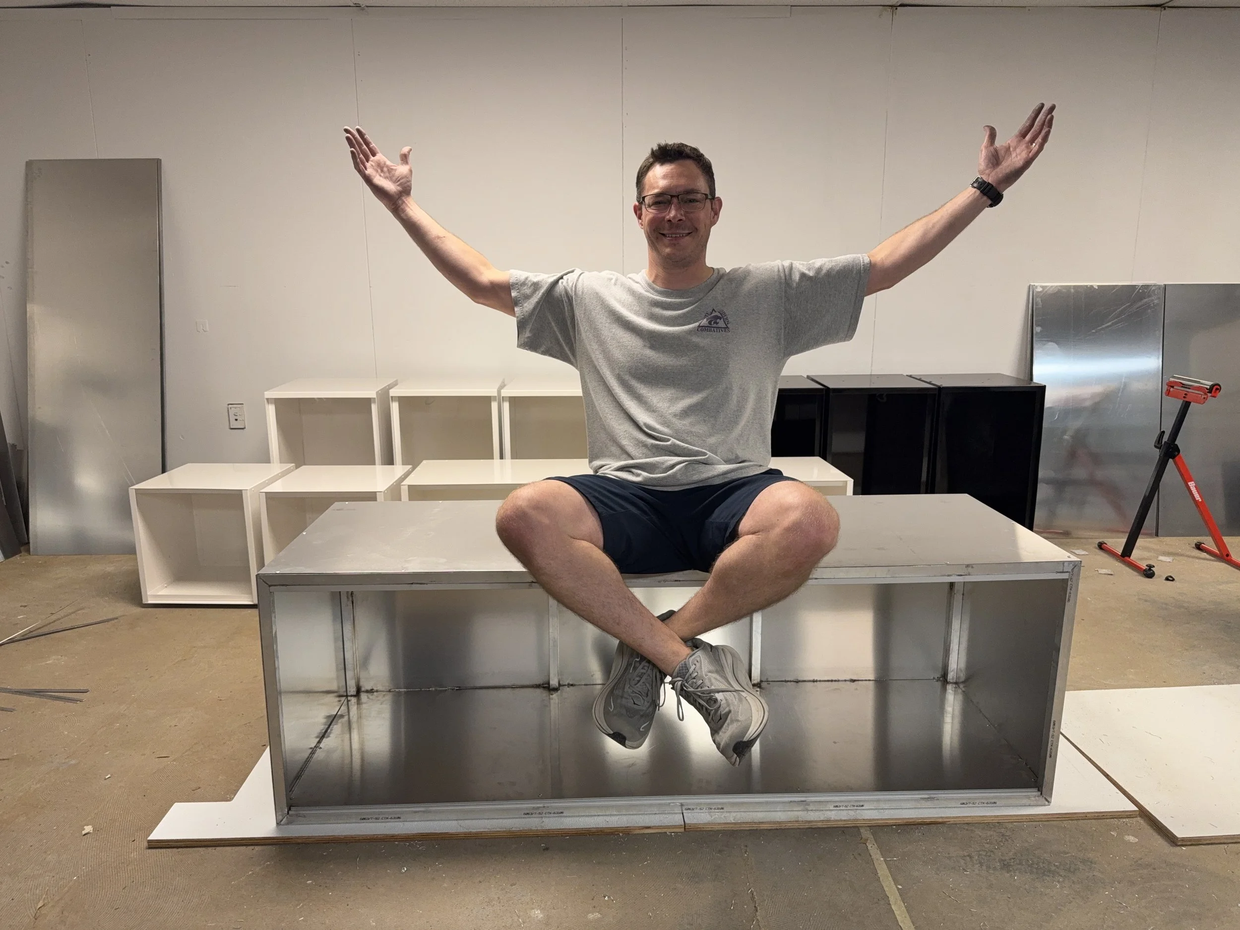 A man, sitting cross-legged on an aluminum reptile enclosure with his arms raised and smiling. The background includes empty white shelving units, and metallic panels, in a room with unfinished flooring.