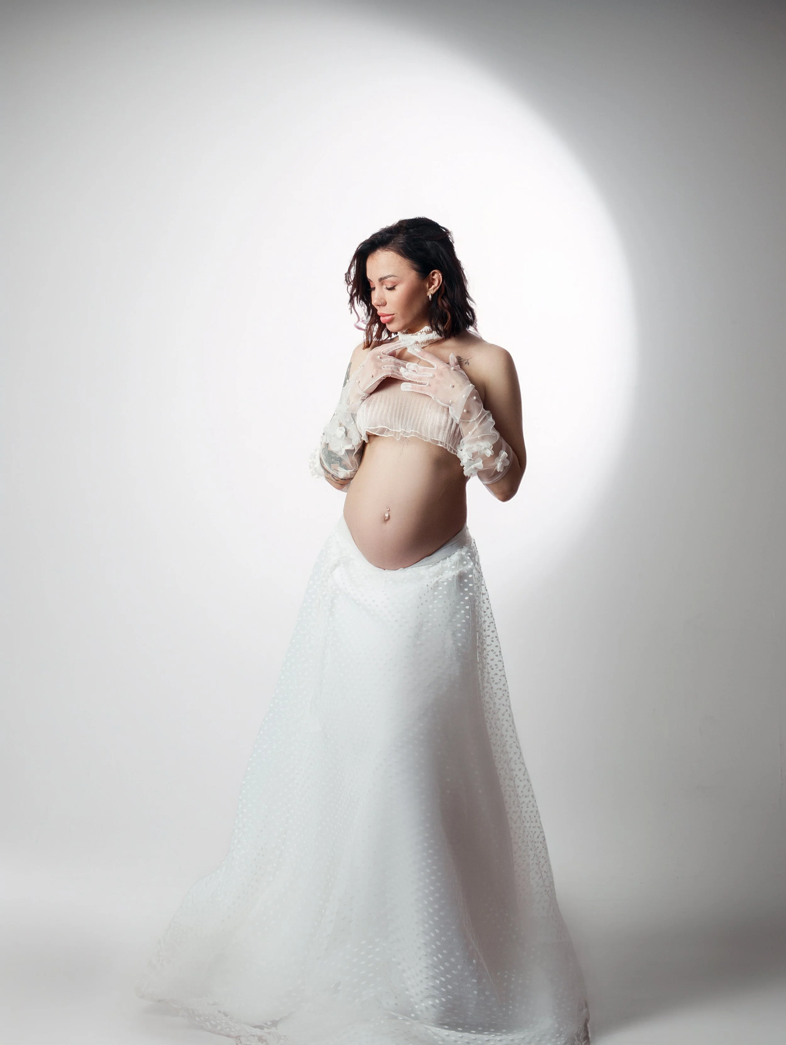 Une femme enceinte portant une robe blanche en dentelle et des gants transparents, se tenant de profil dans une lumière centré sur elle avec un fond blanc.
