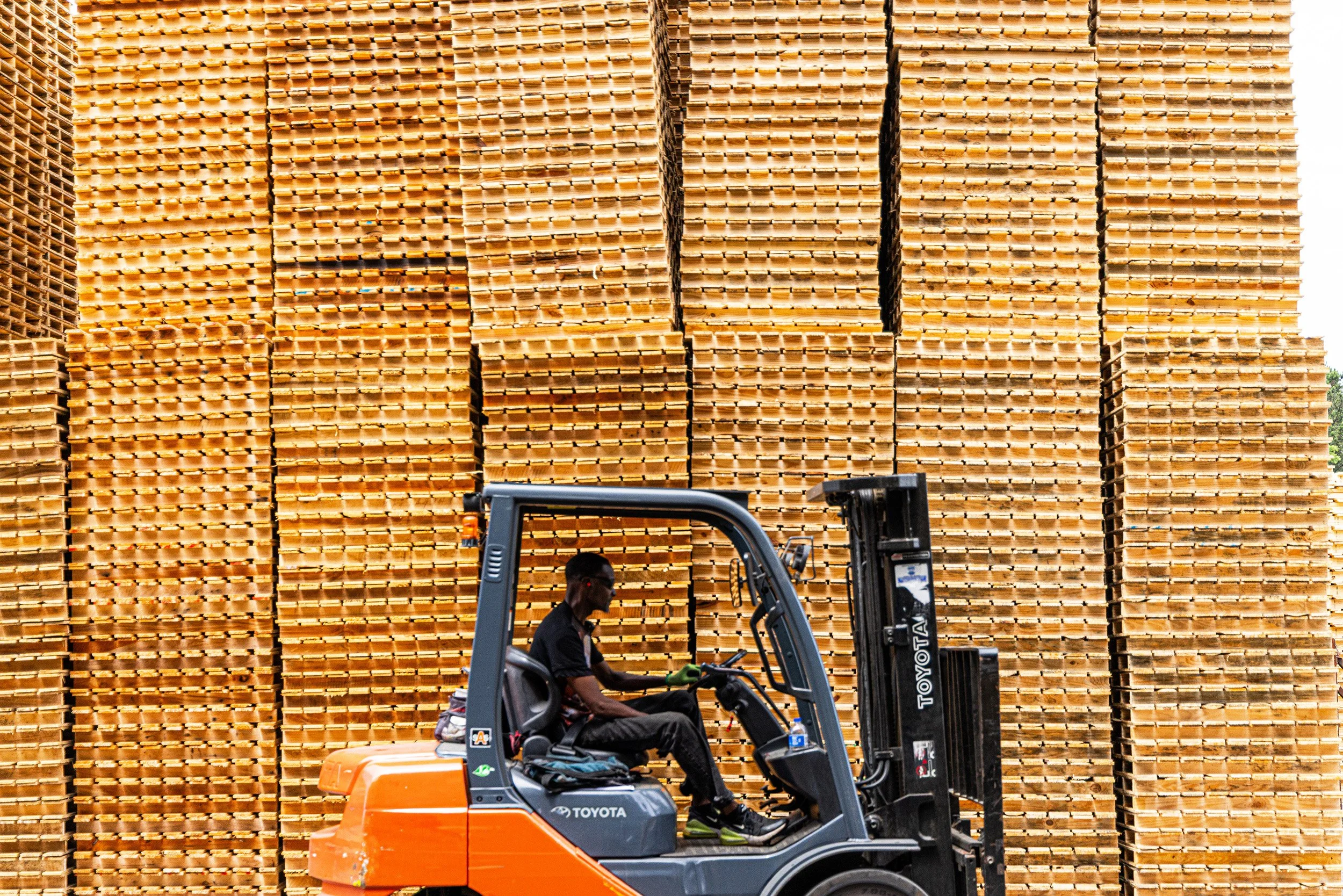 A person operating an orange and gray Toyota forklift in front of a large stack of wooden pallets.