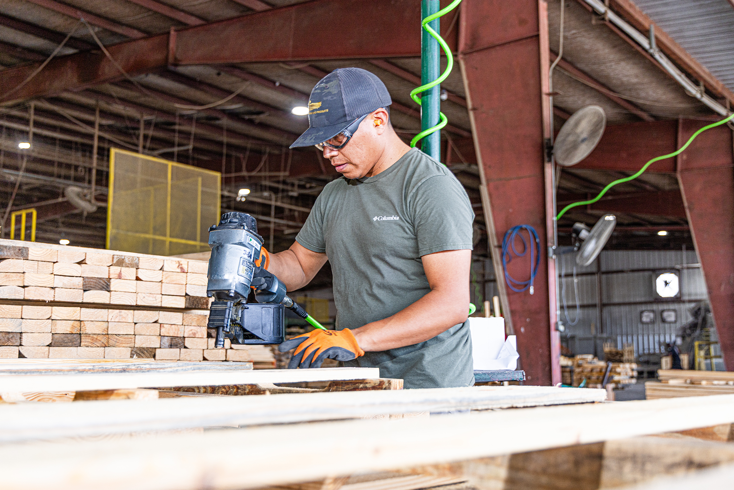 A male worker in a workshop using a nail gun on a stack of wooden planks. He is wearing protective glasses, a baseball cap, and orange gloves.