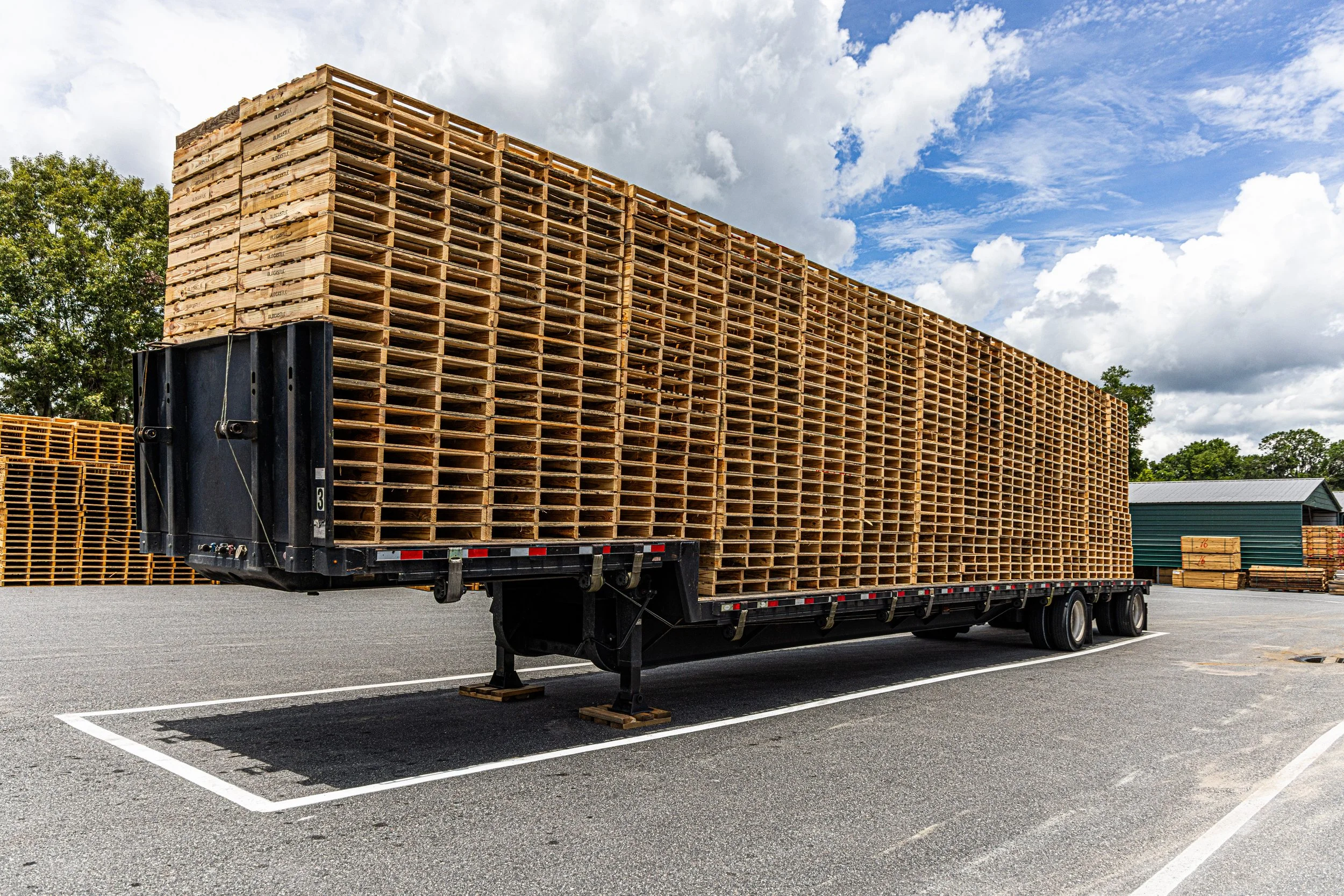 Loaded flatbed truck carrying a large stack of wooden pallets in a parking lot under a partly cloudy sky.