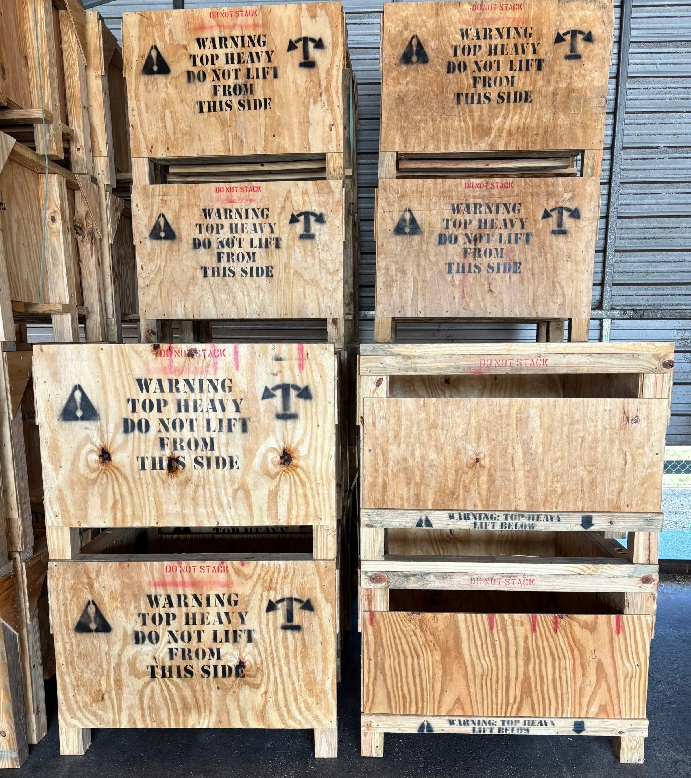 Wooden crates with black spray-painted warning signs stating, 'Warning Top Heavy Do Not Lift From This Side,' with a caution triangle and an axe symbol, on a sidewalk next to a metal wall.