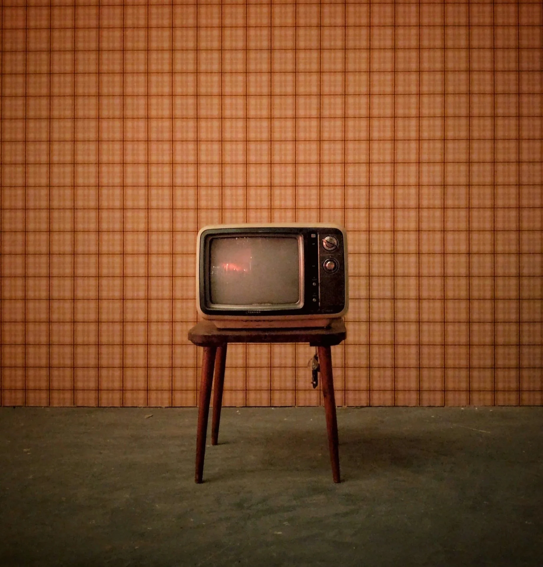An old black-and-white television on a wooden stool in a room with checkered orange wallpaper and a dark floor.