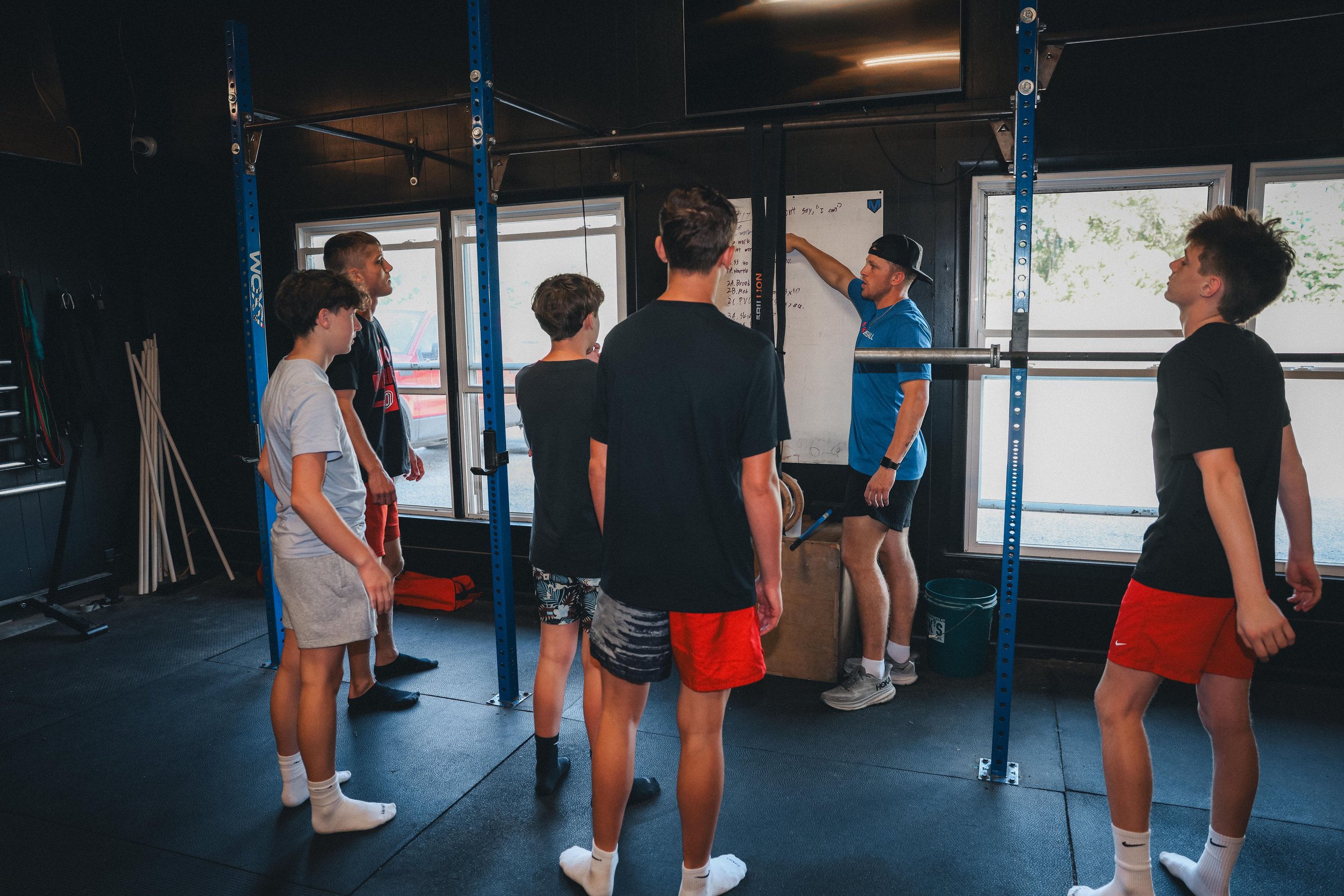 Group of six young people, five males and one female, gathered around a trainer in a gym. The trainer is pointing at a whiteboard as the group listens attentively.