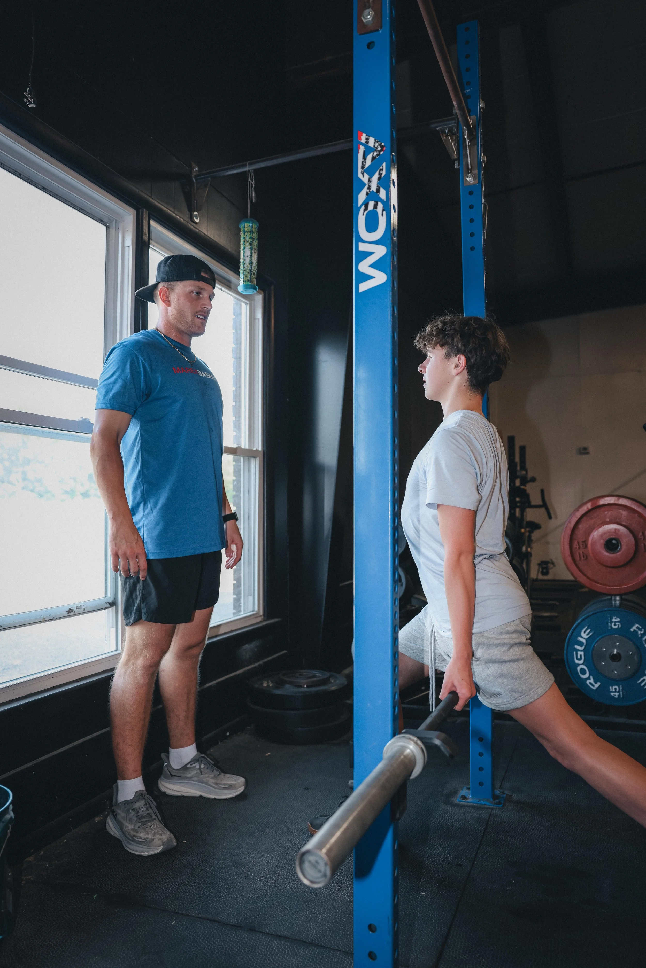 A young man in gray shorts and a white T-shirt with a fitness tracker on his left wrist performing a lunge exercise with a dumbbell in his right hand in a gym. He is facing right, looking forward. An older man in a blue T-shirt, black shorts, and a backwards baseball cap is standing beside him, observing or instructing.