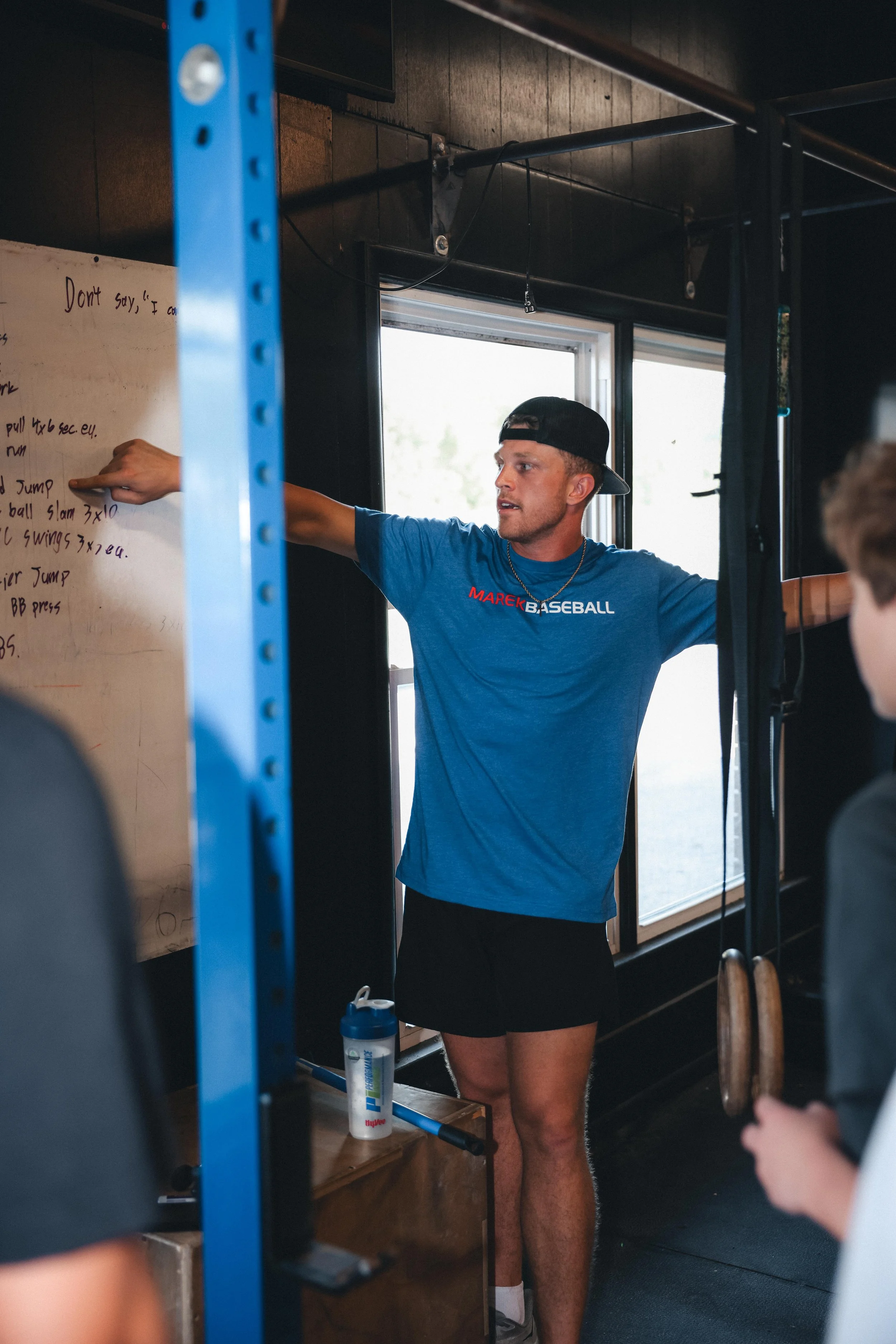 A man in a blue t-shirt and black shorts is standing in a gym, pointing at a whiteboard with workout instructions. He is wearing a backward black cap. The gym has large windows, and workout equipment and a water bottle are visible in the background.
