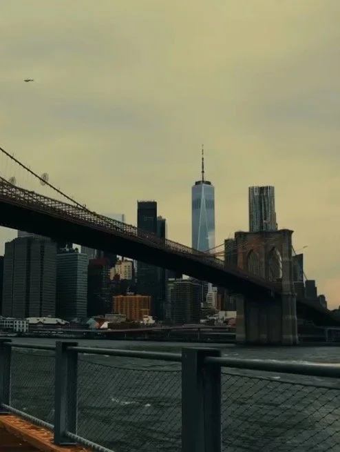 View of New York City skyline with the Brooklyn Bridge in foreground and One World Trade Center in the background, under a cloudy sky.