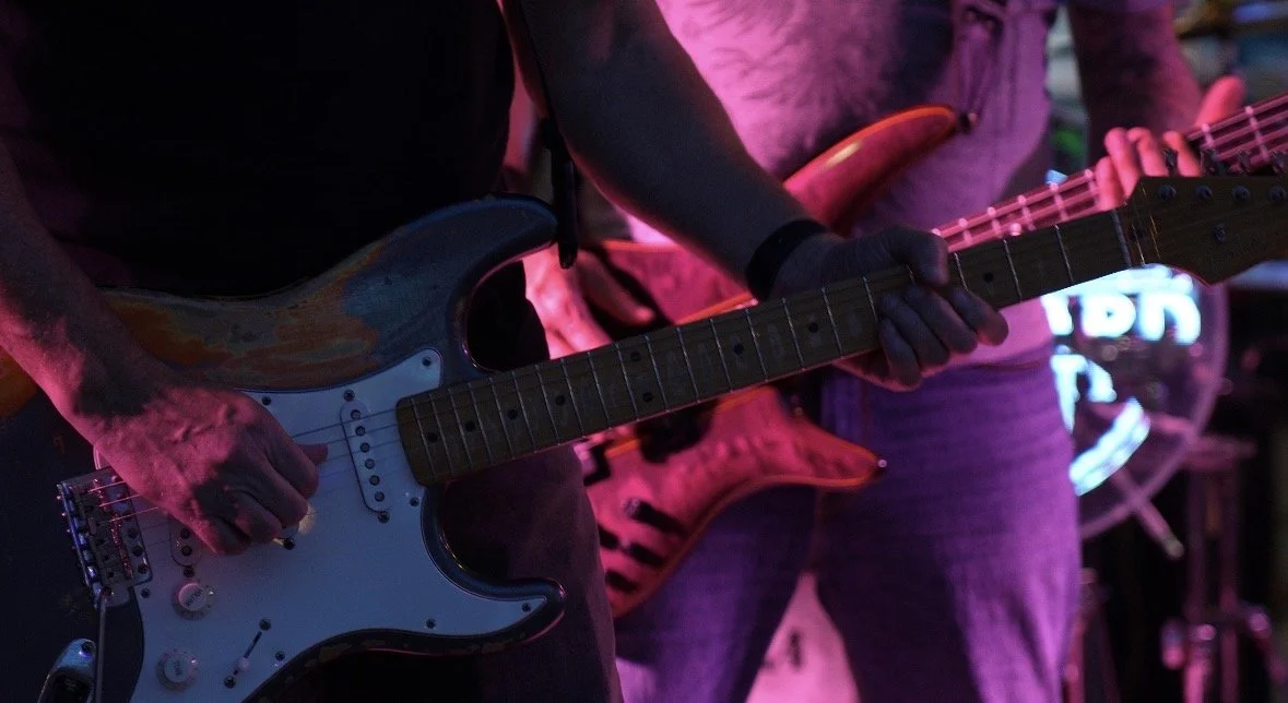 Two musicians playing electric guitars on stage, with purple and pink lighting, at a live music performance.