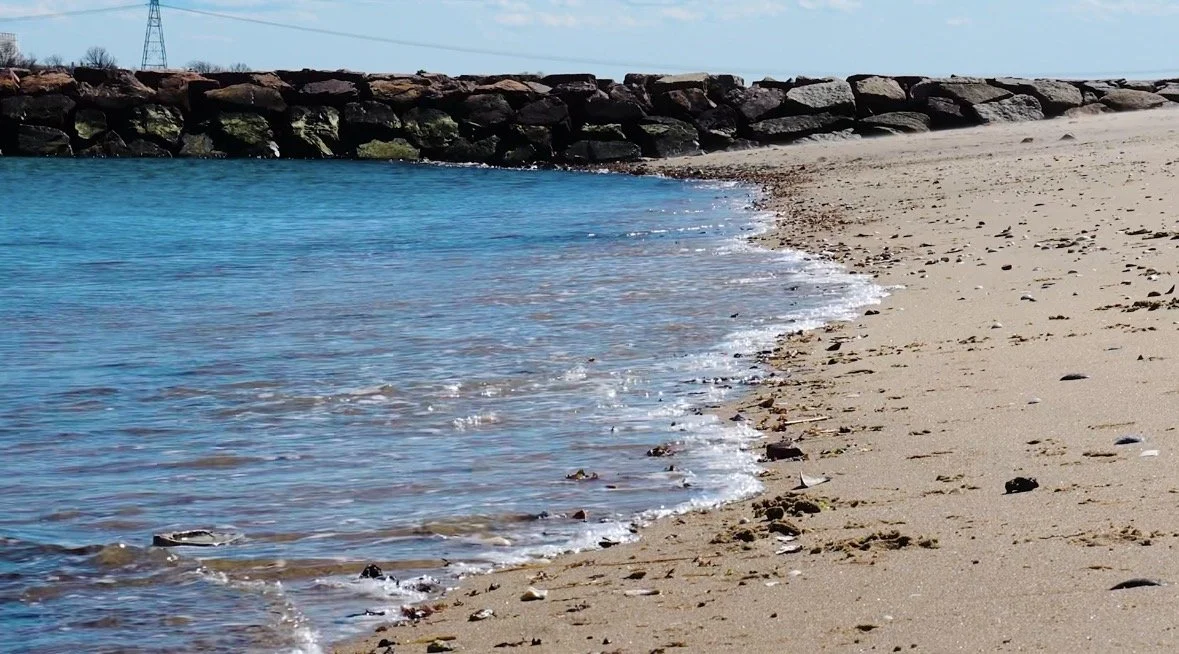 A sandy beach with small rocks and shells along the shore, gentle waves lapping against the sand, a stone breakwater in the background, and clear blue sky above.
