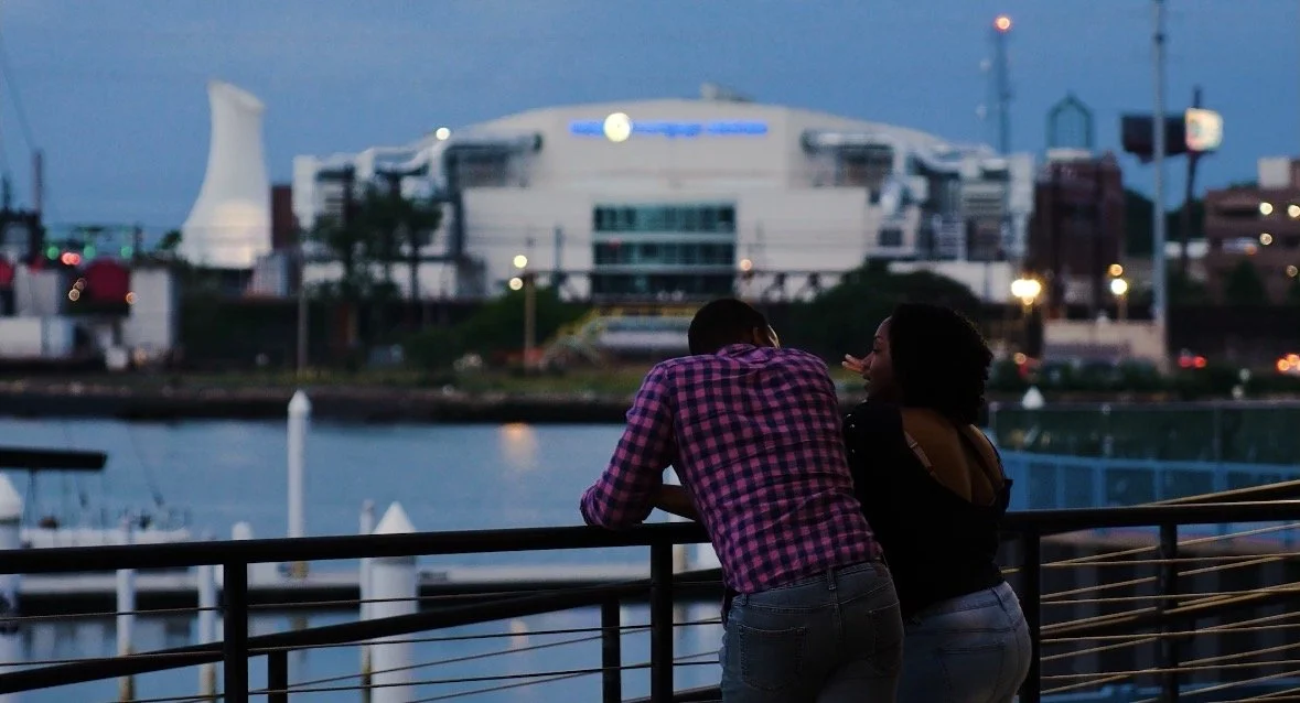 A couple leaning on a railing by the water during dusk, with a cityscape in the background.