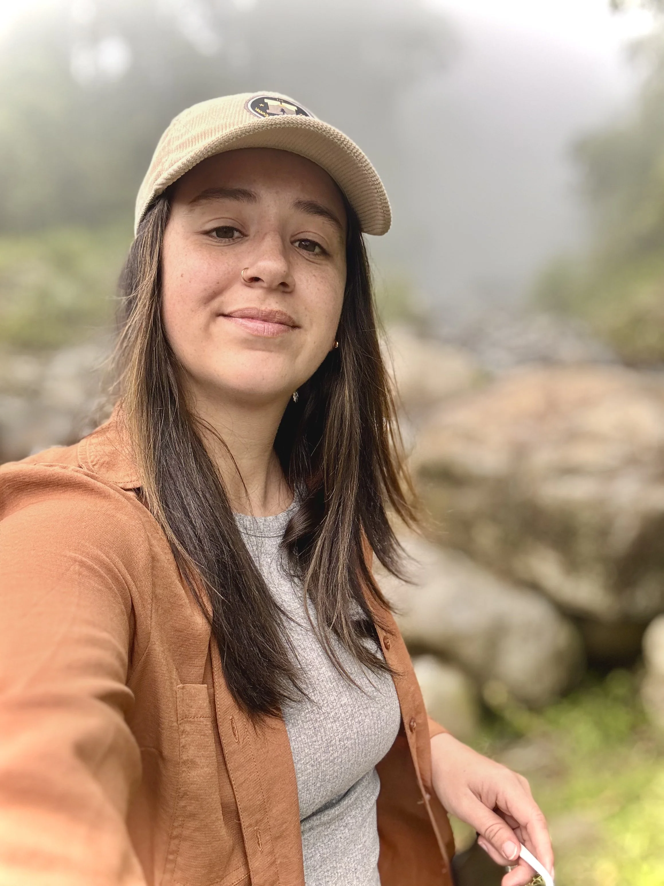 A woman with long brown hair, wearing a beige cap with a patch and a nose ring, taking a selfie in Costa Rica with blurred greenery and rocks in the background.