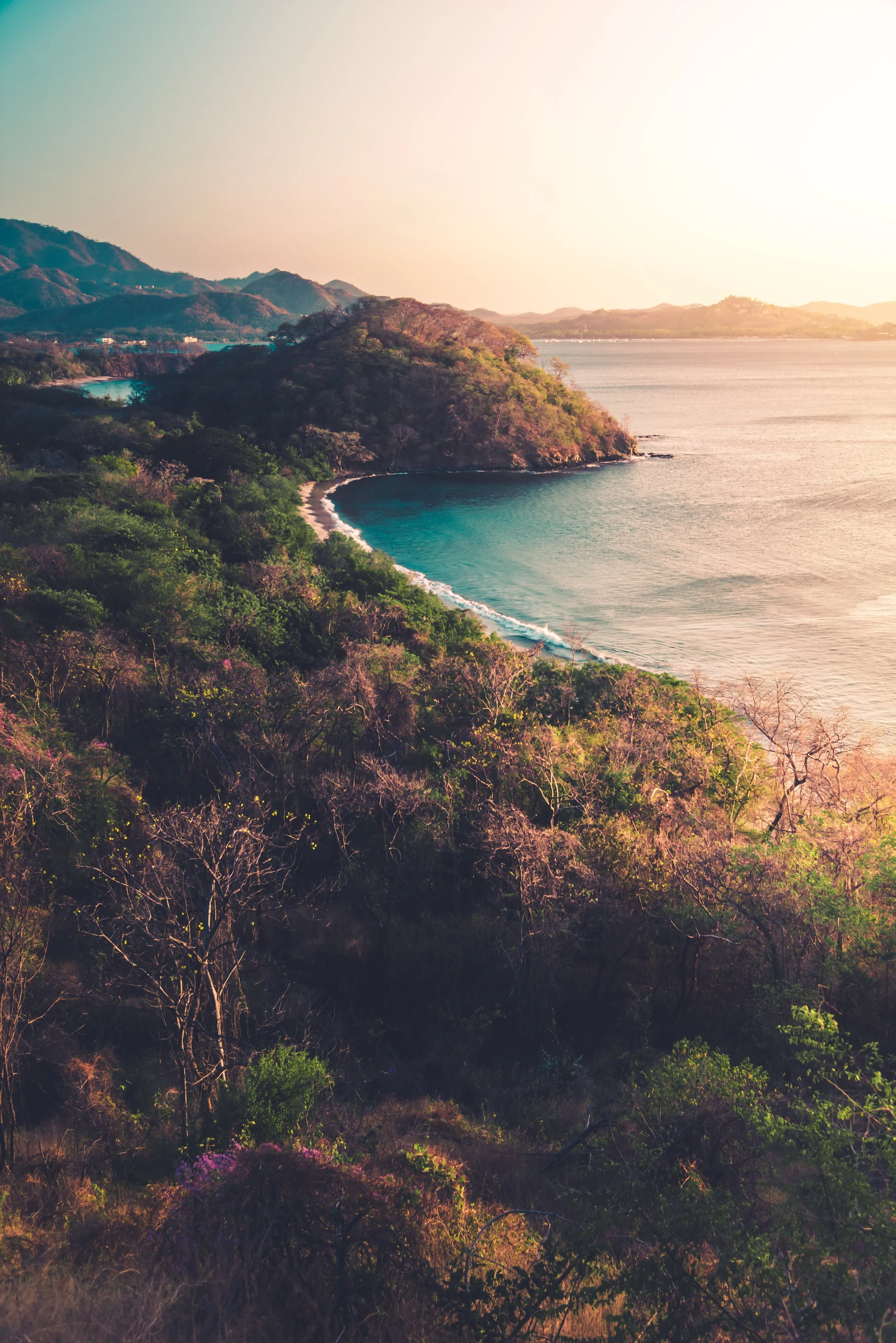 Sunset over a coastal Costa Rica view with lush greenery on a hillside, calm water, and distant mountains.