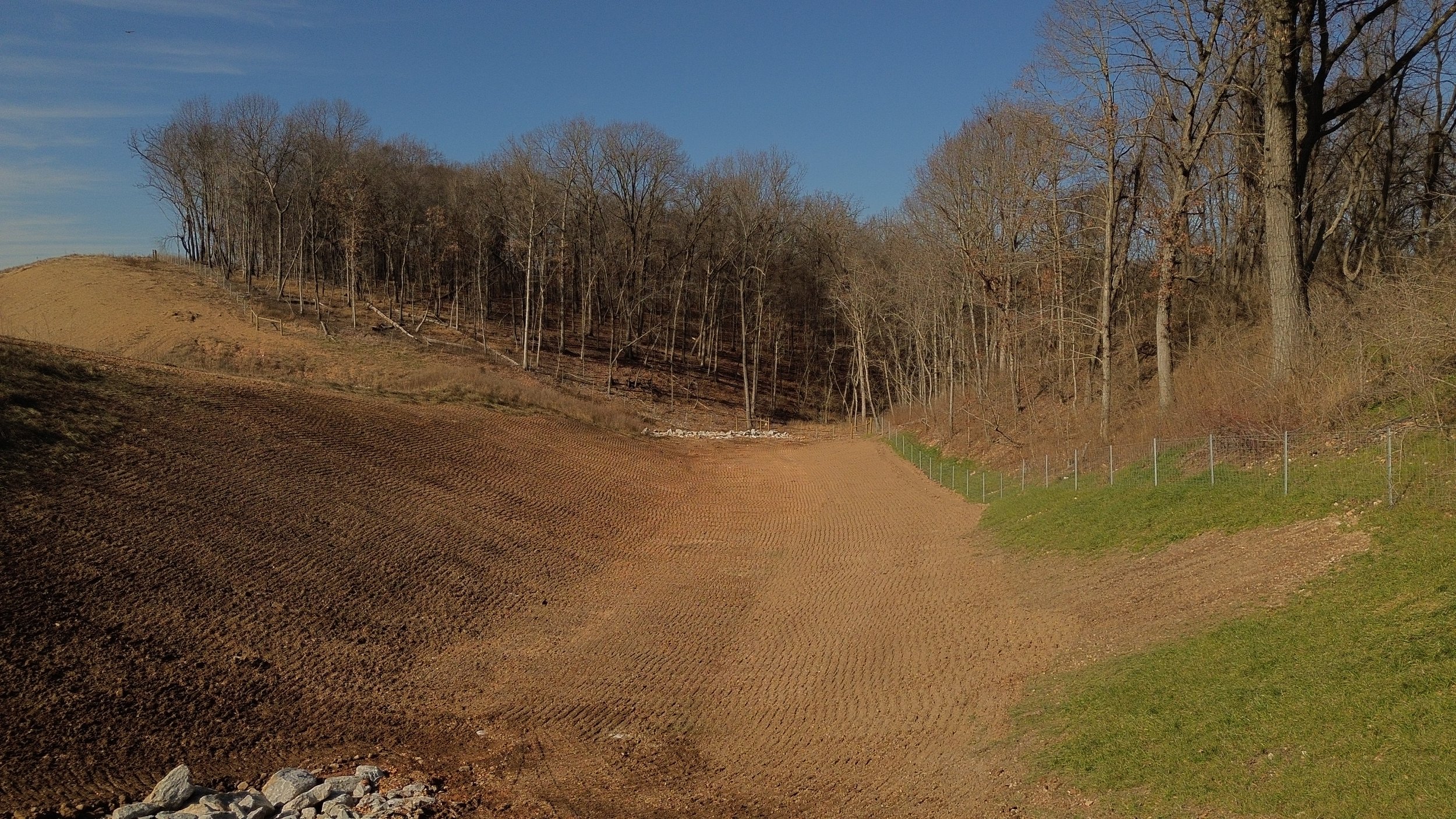 A rural landscape with a dirt field in the foreground, a slope with a fence on the left, and a wooded hillside with leafless trees under a clear blue sky in the background.