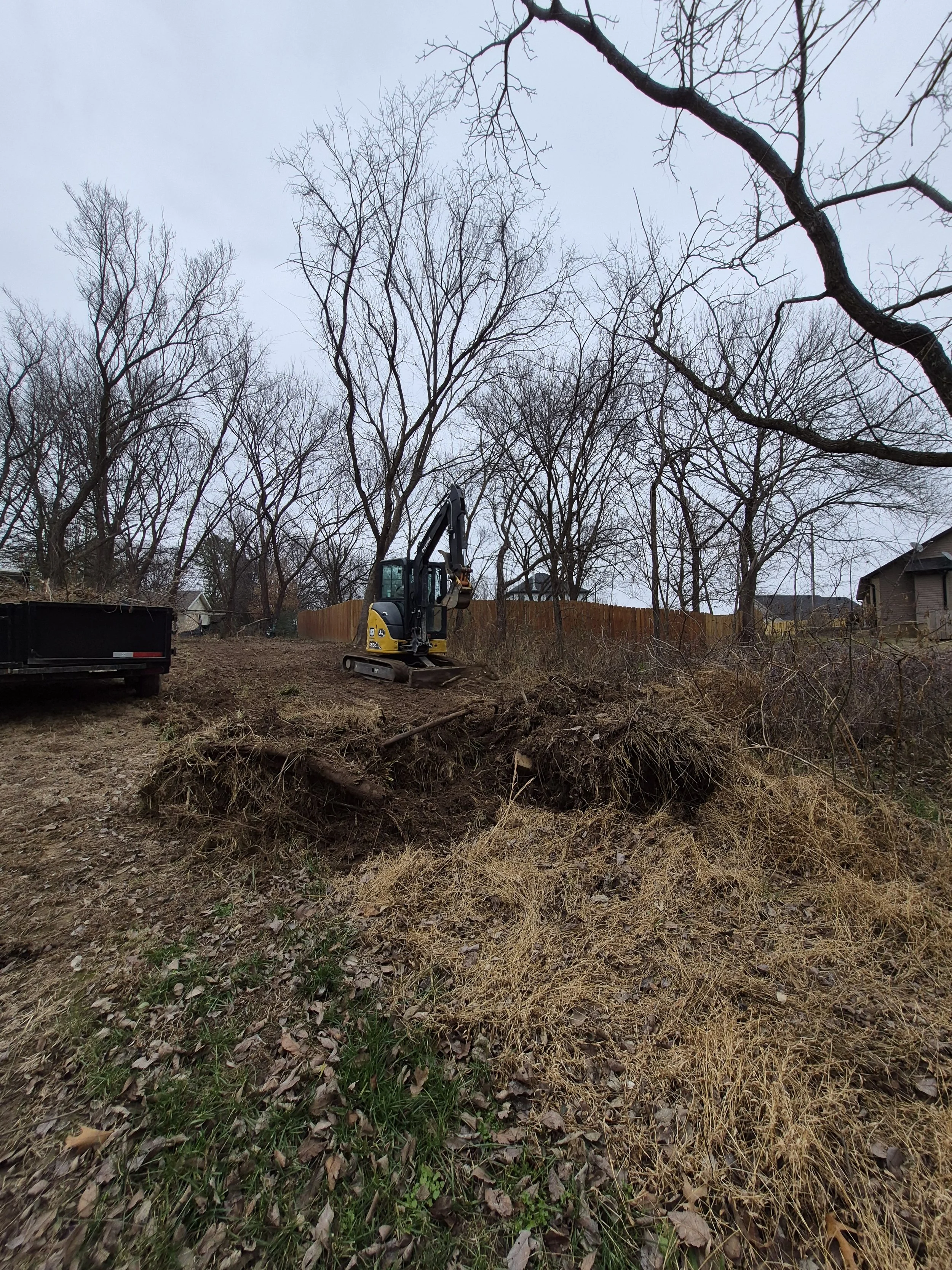 A small excavator on a dirt lot, surrounded by leafless trees and a wooden fence.