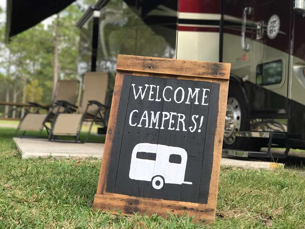 A wooden sign with the words "Welcome Campers!" and a drawing of a camper trailer, placed outside near a recreational vehicle and lounge chairs on a grassy area.