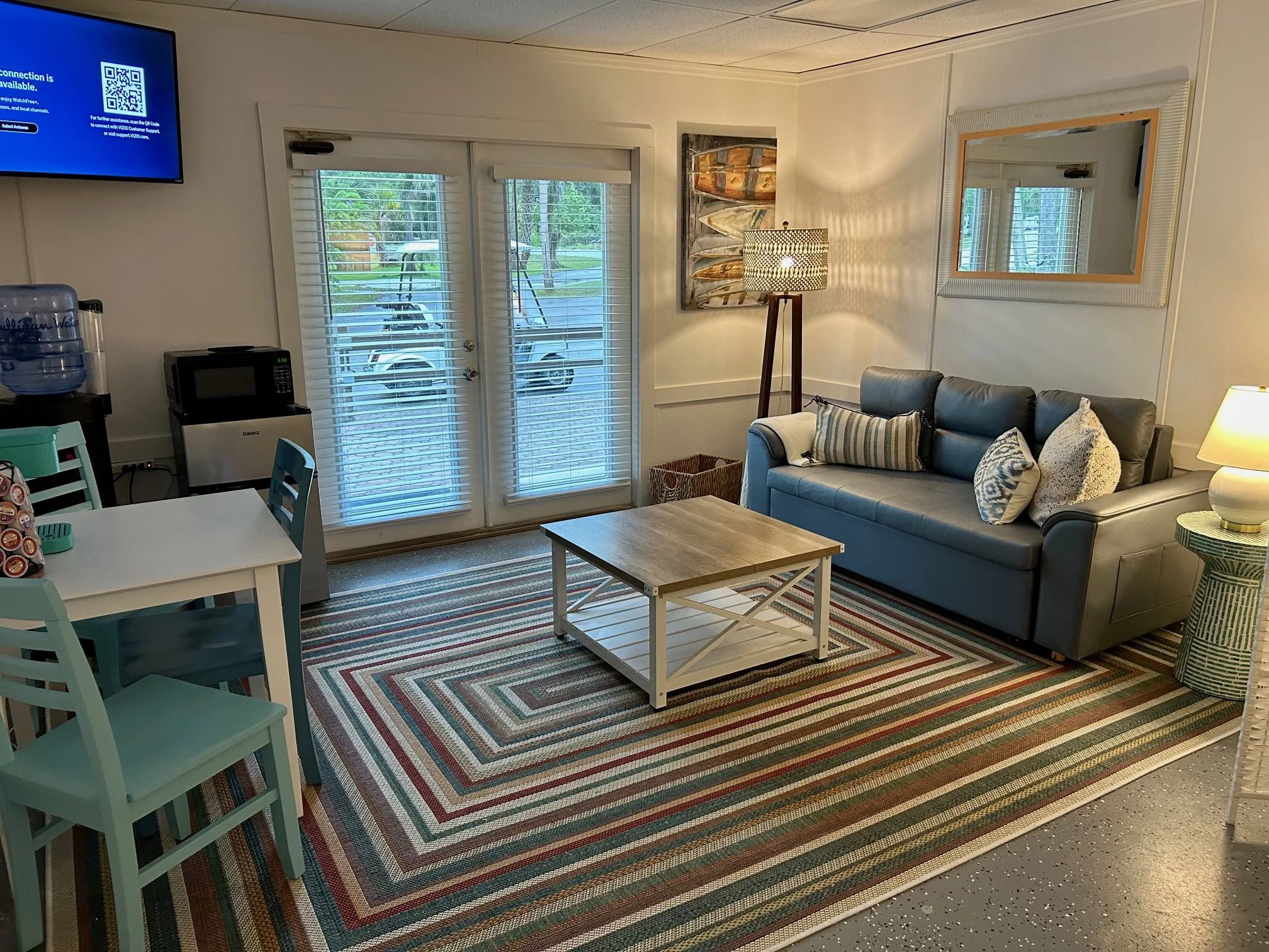 New Smyrna Beach RV Park suite with a striped area rug, a gray couch with pillows, a wooden coffee table, a floor lamp, and artwork of fish above the couch. Sliding glass doors with blinds lead outside to a porch with chairs and greenery.