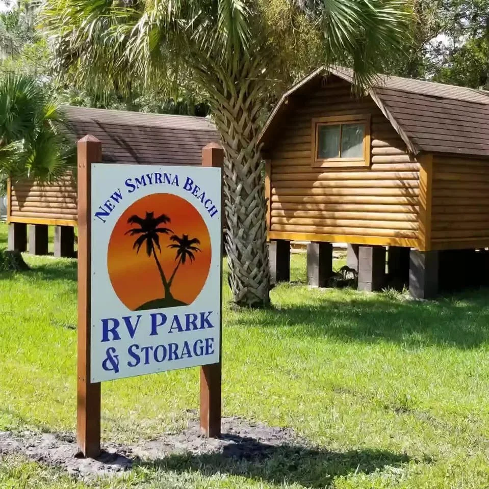 A sign for New Smyrna Beach RV Park & Storage with palm trees and a sunset graphic, in front of wooden cabins on stilts and surrounded by grass and palm trees.