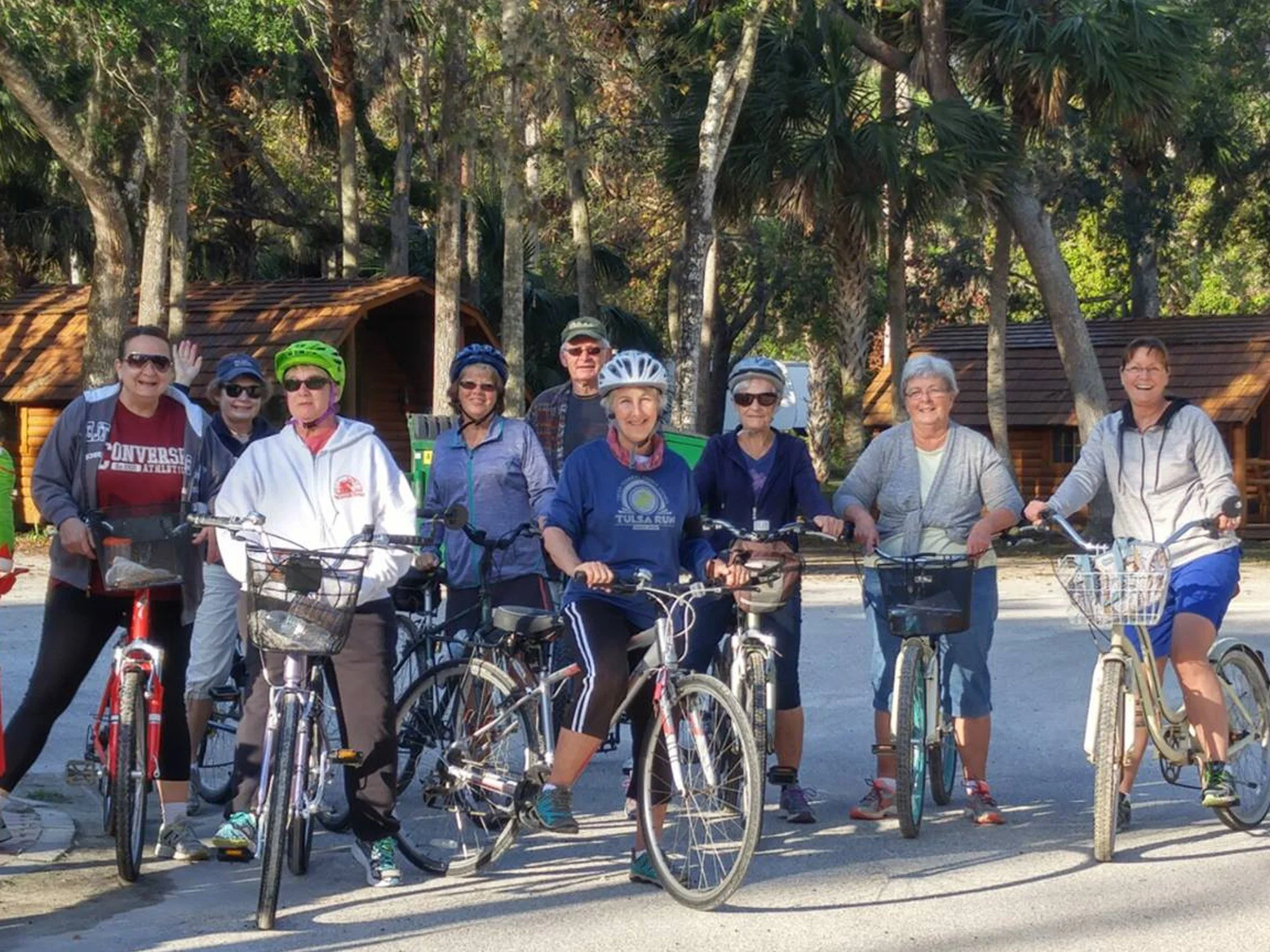A group of elderly people with bicycles standing outdoors in a park area surrounded by trees and wooden cabins, smiling and wearing helmets.