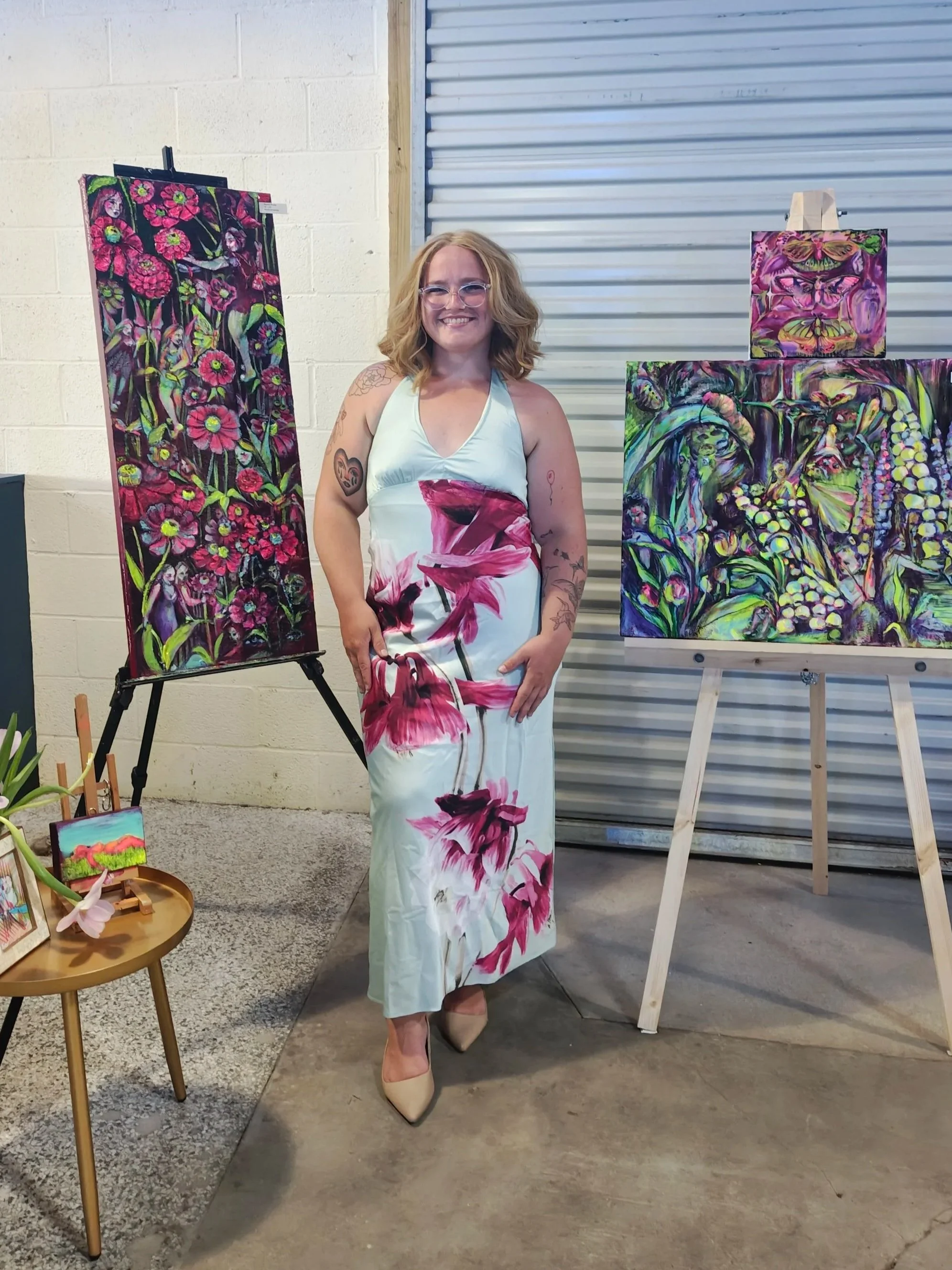 A woman in a white dress with pink floral patterns, wearing beige high heels and glasses, standing in front of several colorful artwork paintings at an art gallery or exhibition.