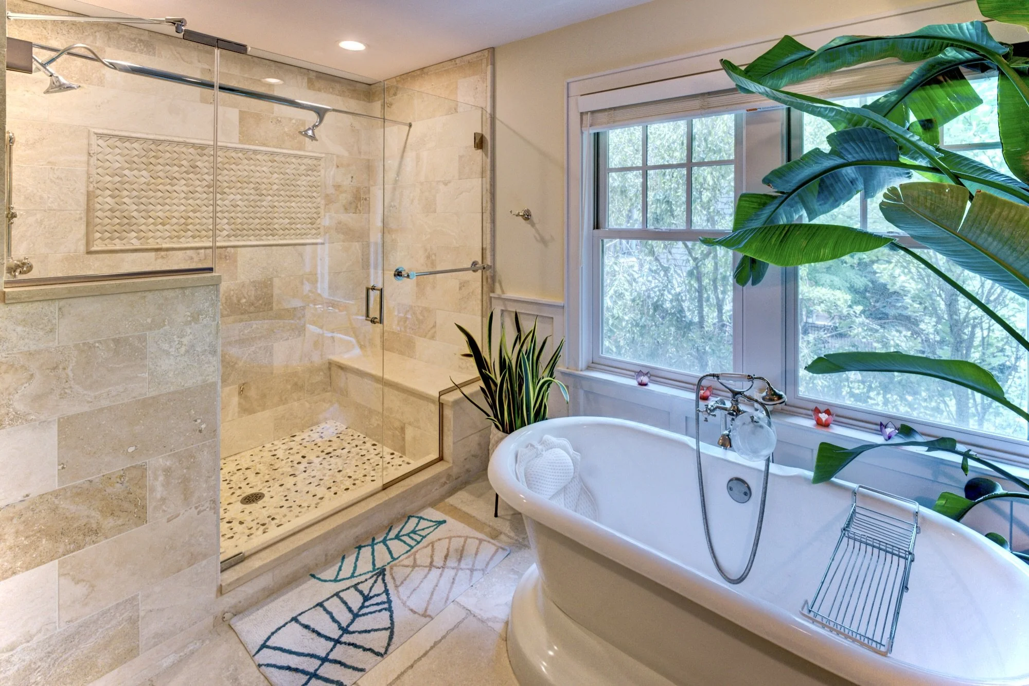 A bathroom with a glass-enclosed shower on the left, featuring beige tiles and a built-in niche. To the right, there is a large window with greenery outside, and a white bathtub with a silver faucet and a small rack on the right side. The bathroom ha