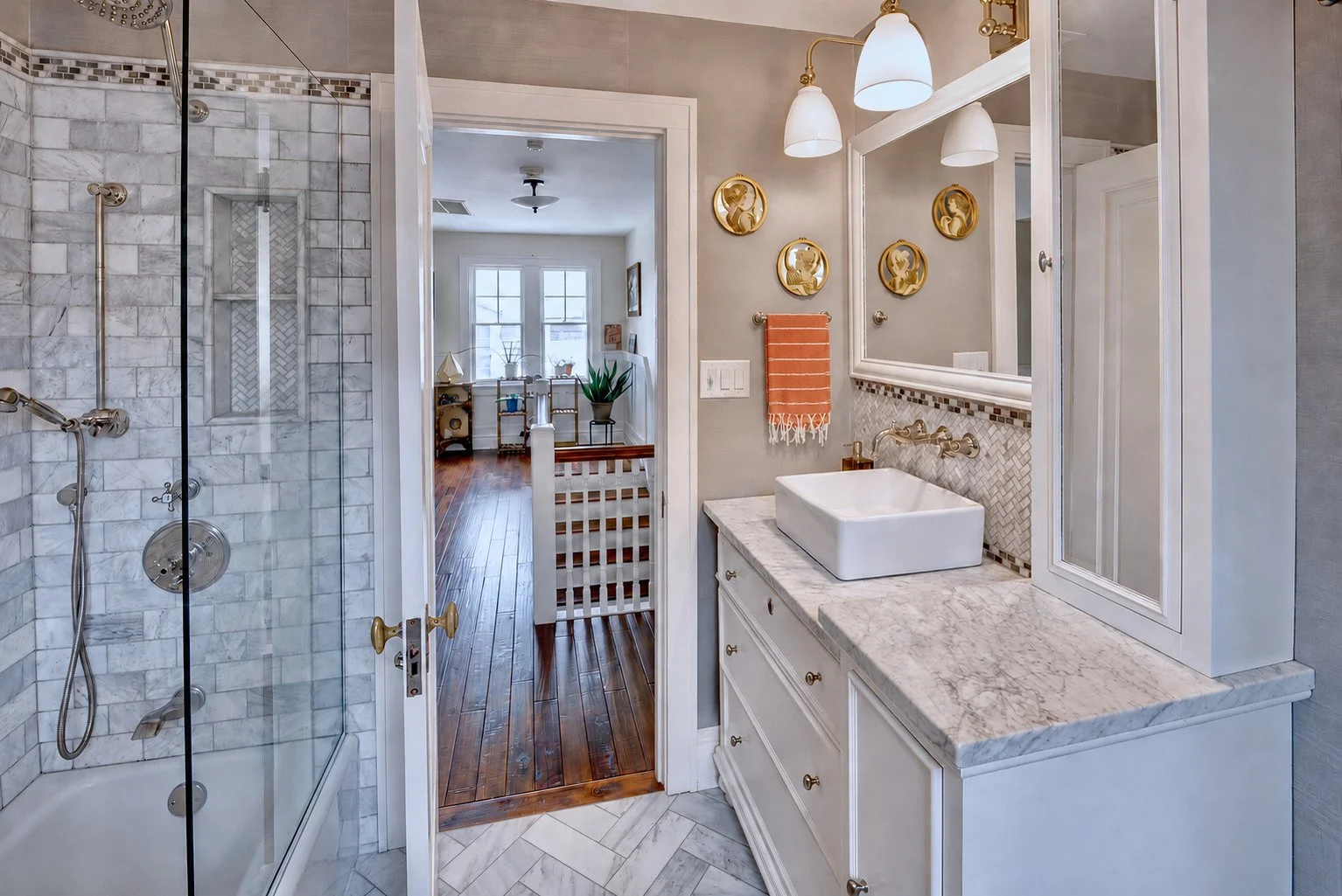 A view of a bathroom with a glass-enclosed shower on the left, a marble countertop with a vessel sink on the right, and decorative wall art. The bathroom opens into a brightly lit room with wooden floors and windows.